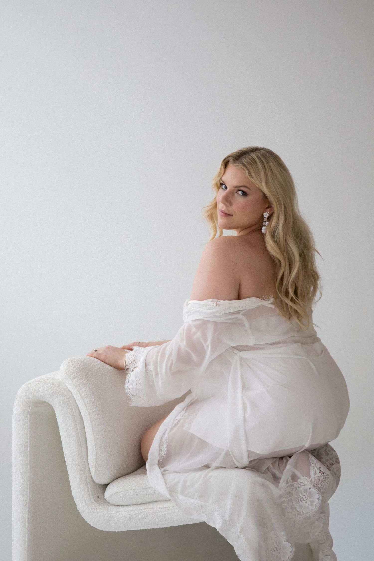 A blonde woman in a white off-shoulder gown sits elegantly on a white chair, looking over her shoulder in a studio.