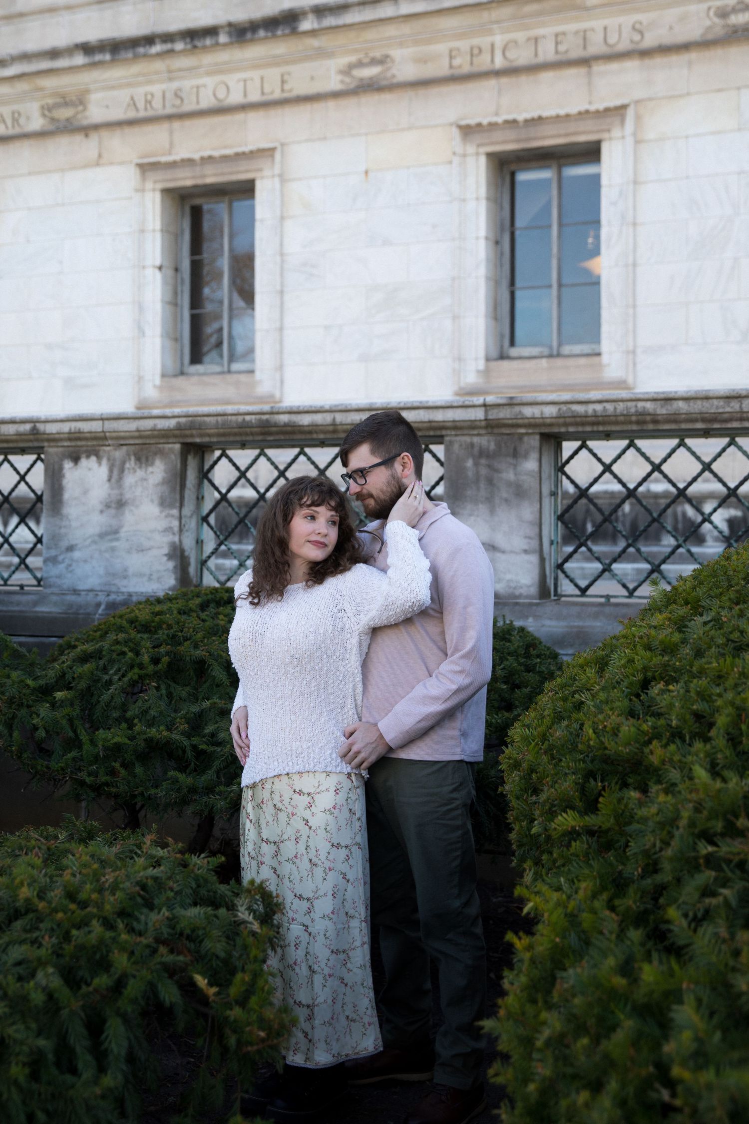 Couple embracing in front of a classical building engraved with Aristotle and Epictetus, surrounded by trimmed hedges.