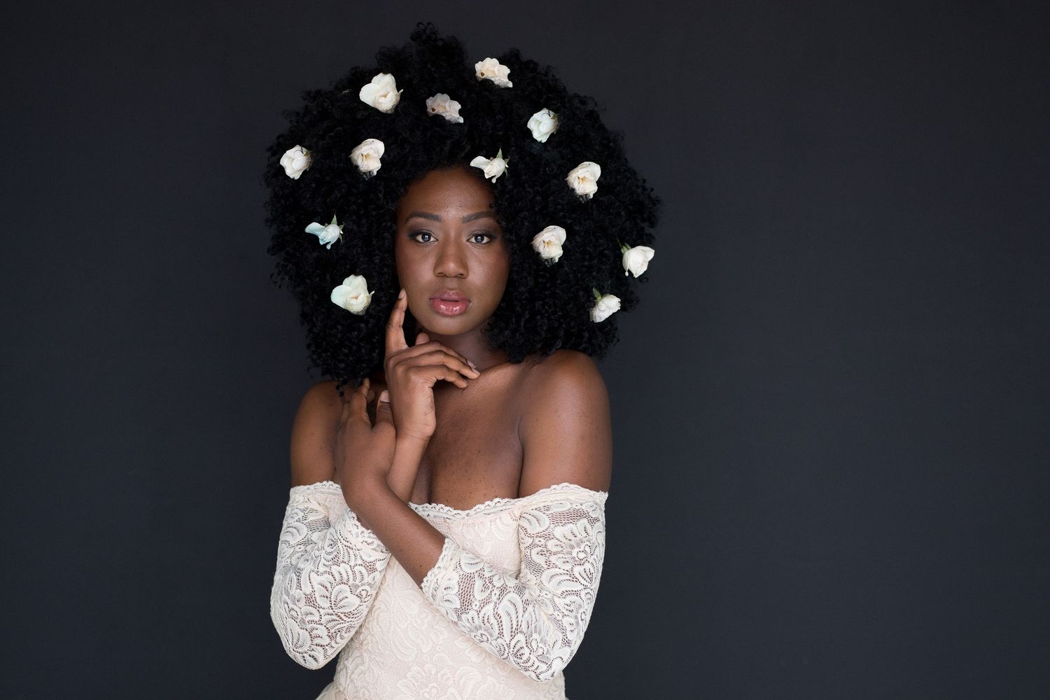 Black woman in white lace off-shoulder gown with white flowers in natural afro hair poses against dark background.