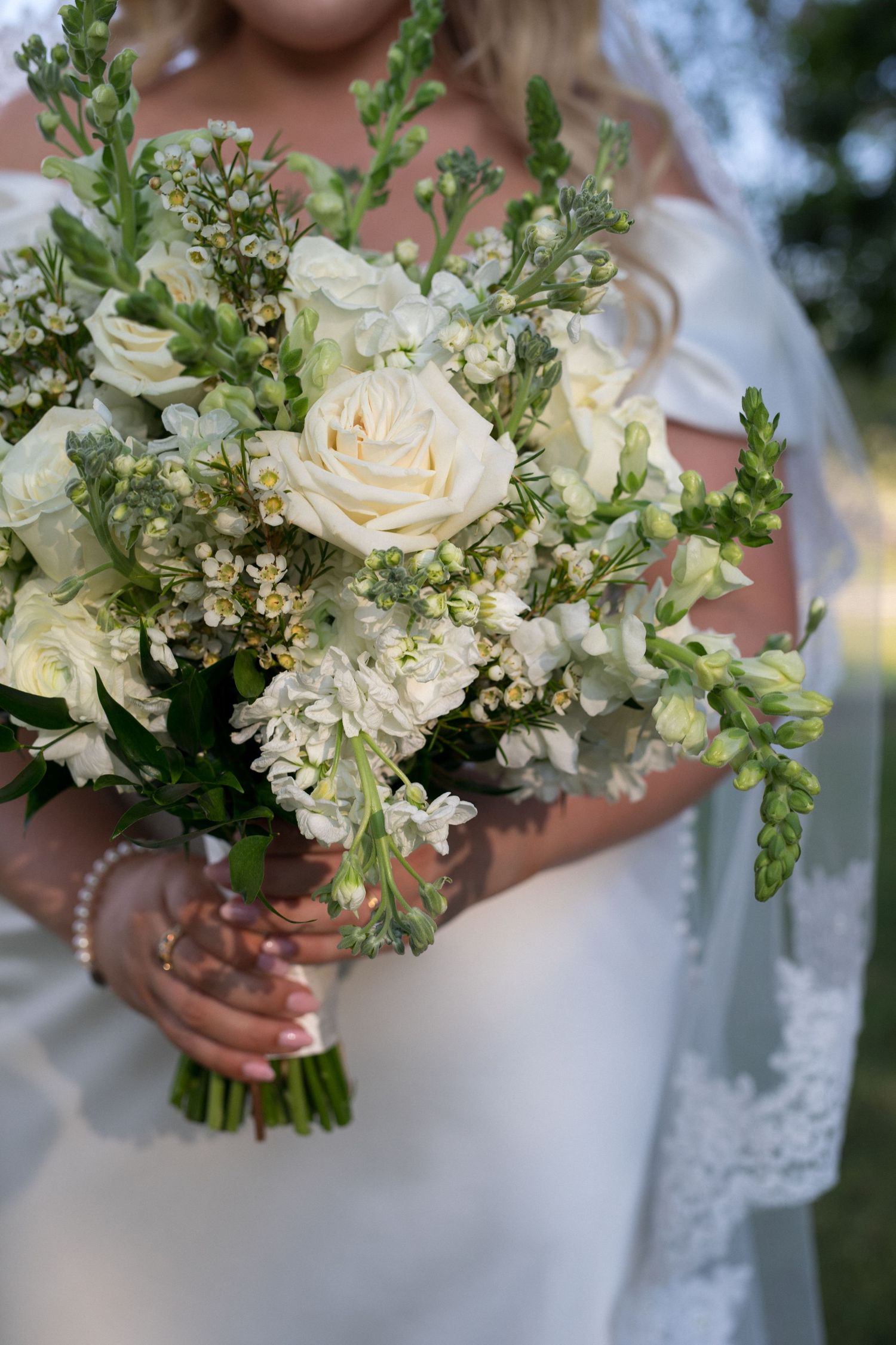 A delicate white wedding bouquet featuring cream roses, carnations and greenery held against a white dress.