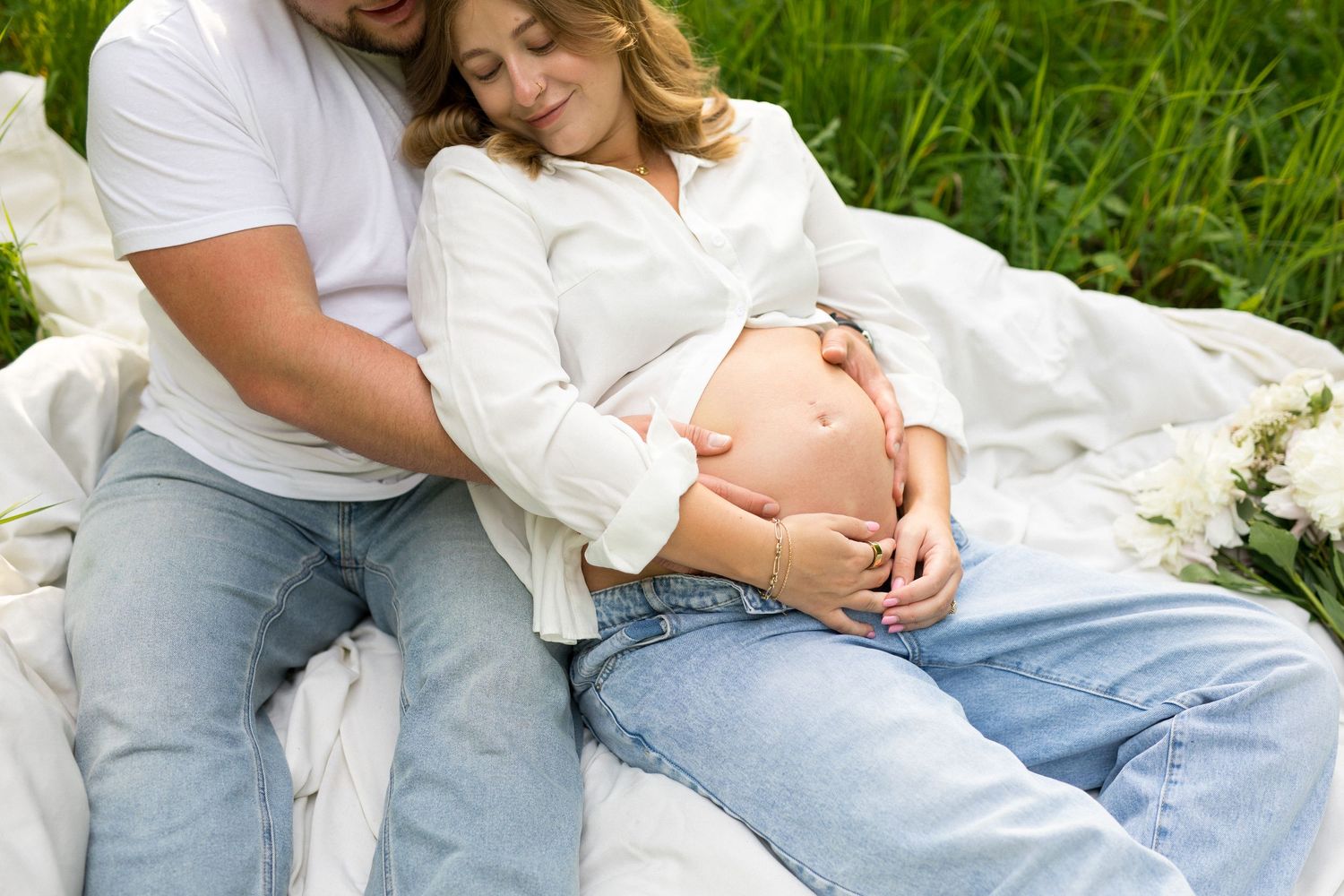 Couple sitting on blanket in grass, pregnant woman in white shirt and jeans cradling baby bump with partner.