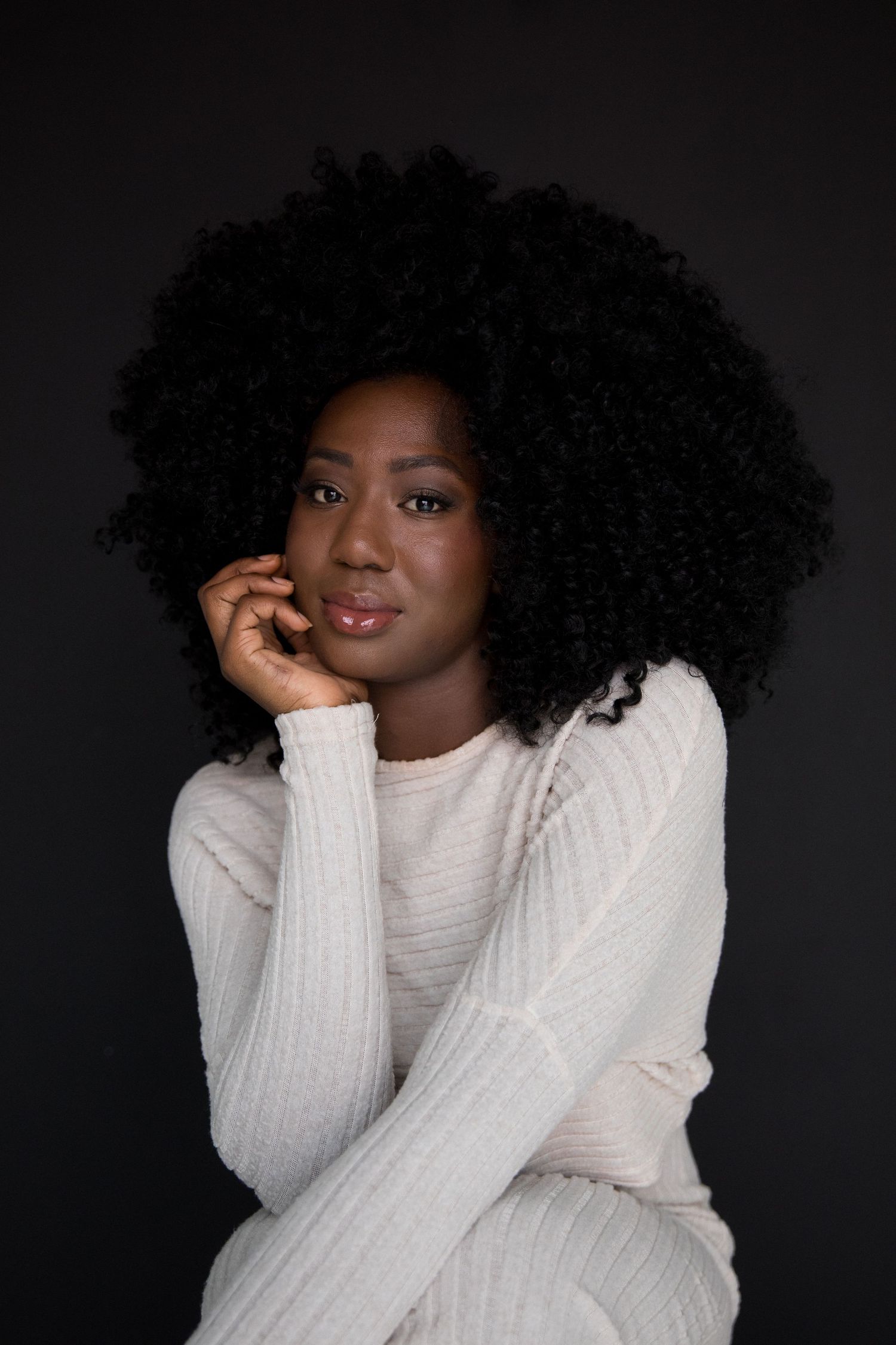 Young Black woman with natural afro hair wearing a white knit sweater, posing thoughtfully against dark background.