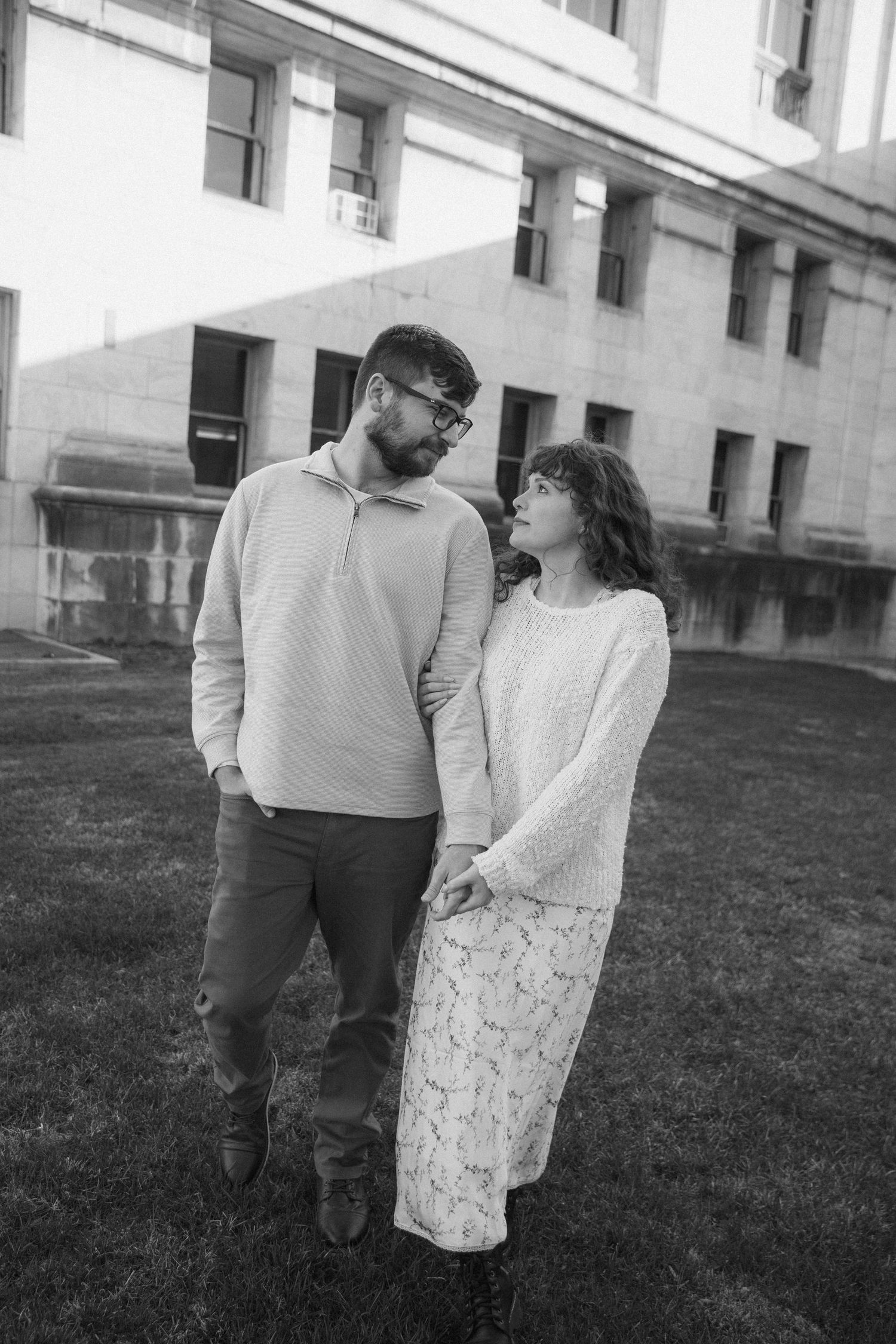 Black and white photo of a couple holding hands and gazing at each other outdoors near a building.
