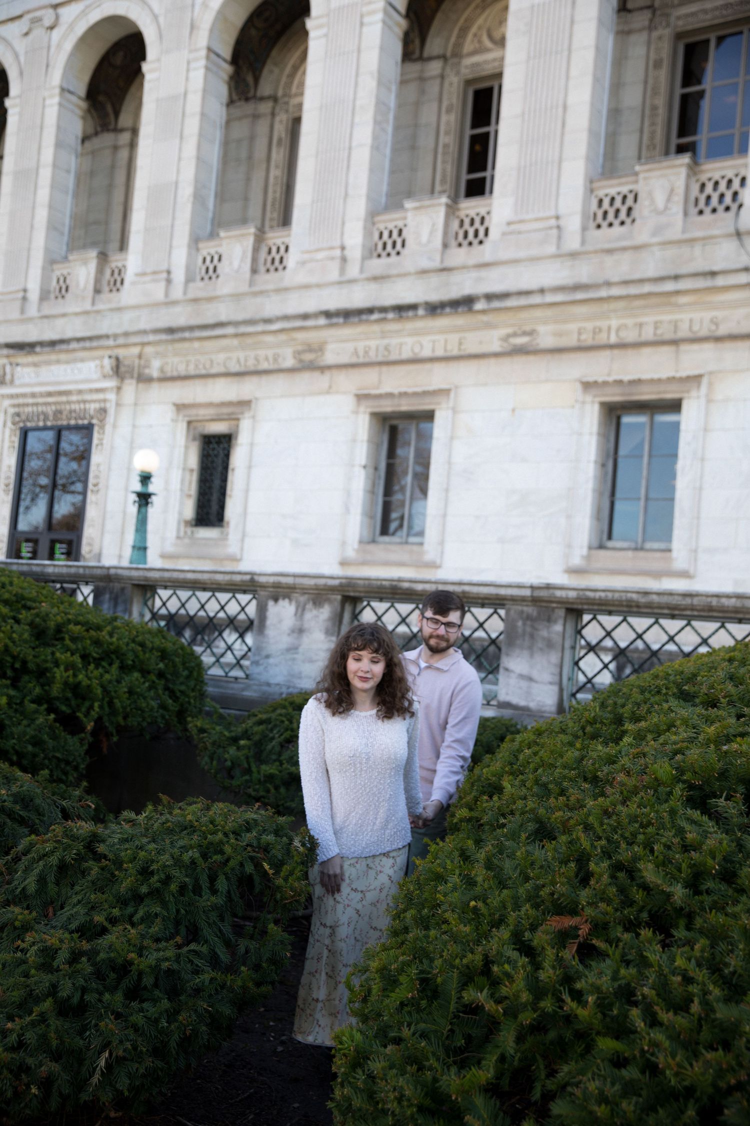 Couple posing in front of a grand neoclassical building surrounded by manicured hedges.