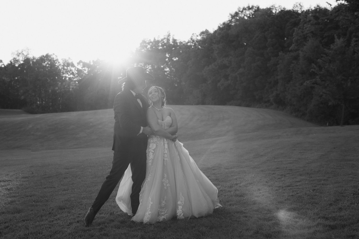 A couple in formal wedding attire dances together in a sunlit meadow at sunset.