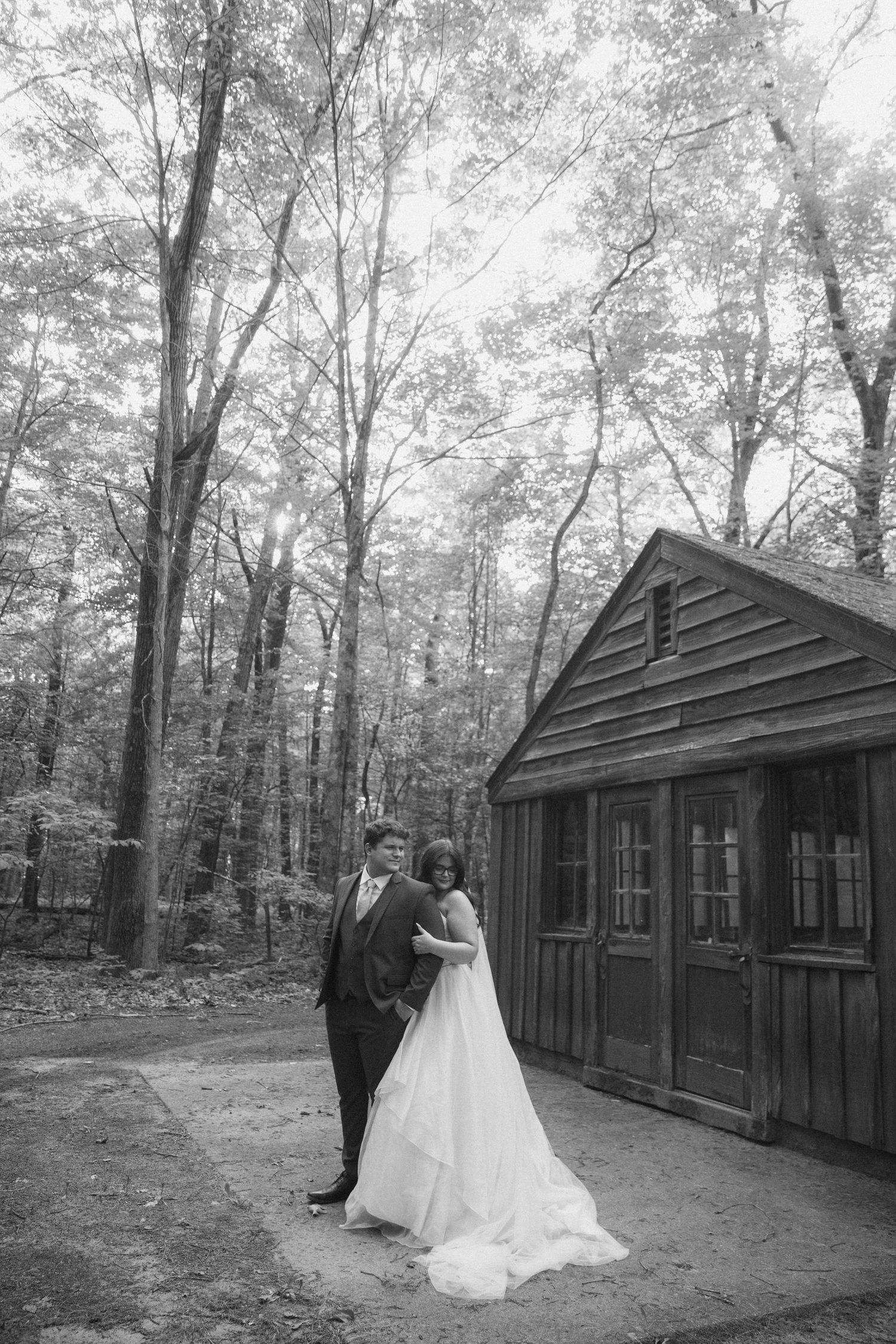 Black and white photo of a bride and groom embracing near a rustic wooden cabin surrounded by tall trees.