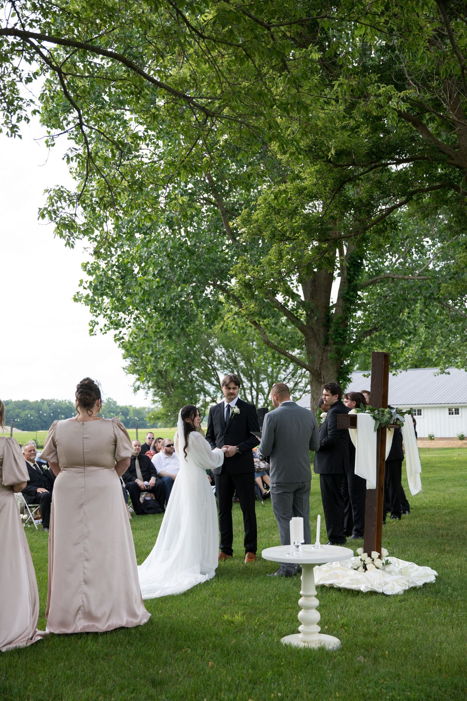 Outdoor wedding ceremony on green lawn under large trees with bride, groom, officiant, and wedding party gathered near a wooden cross.