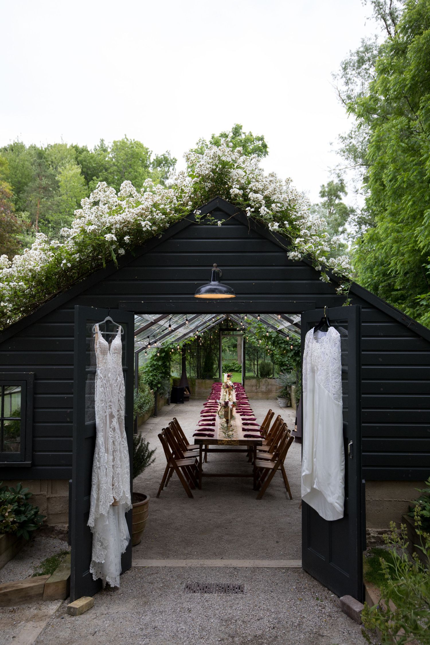 White wedding dresses hang on display in a rustic black wooden pavilion with rocking chairs and climbing vines.