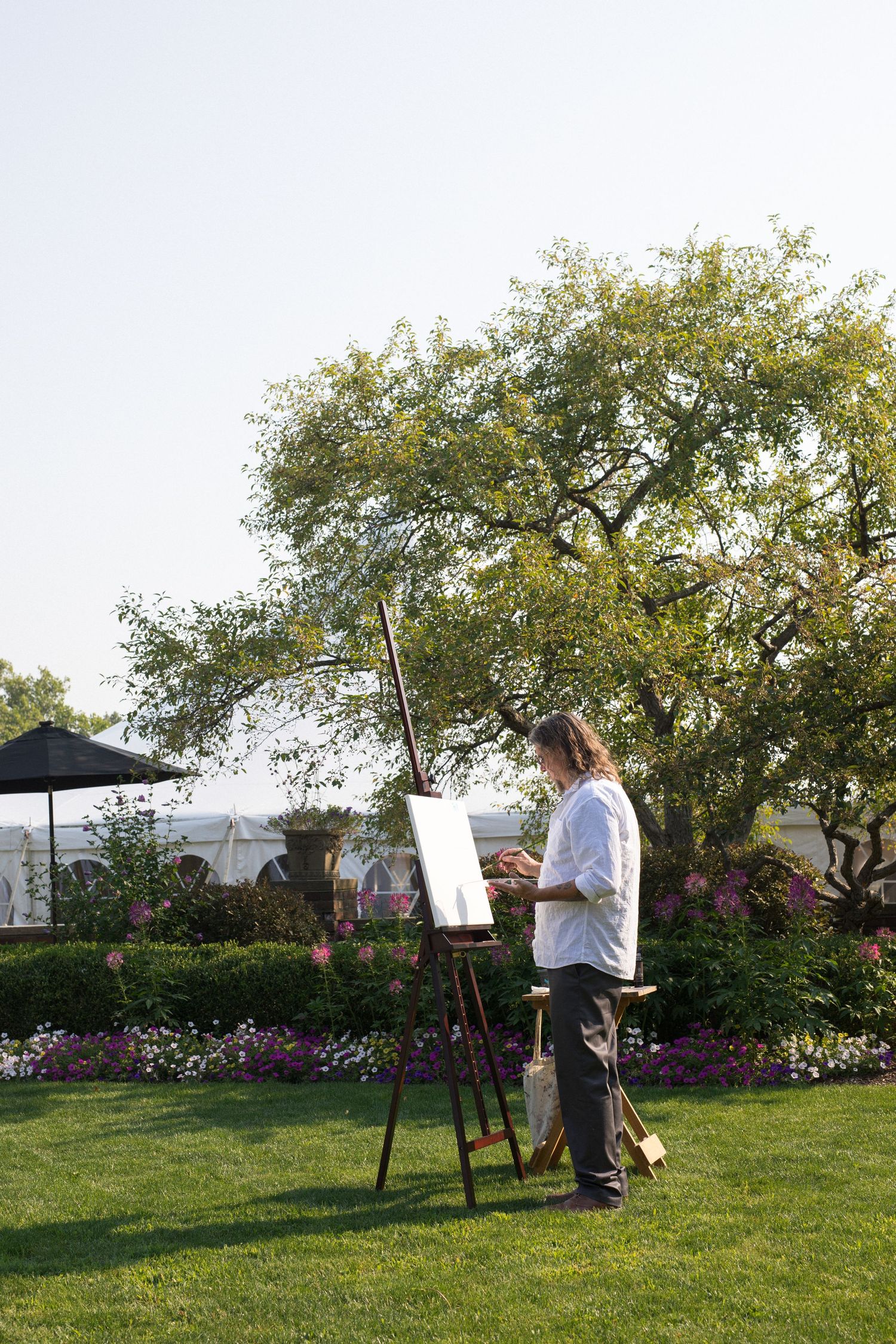Artist paints on easel outdoors in garden setting with blooming flowers and trees.