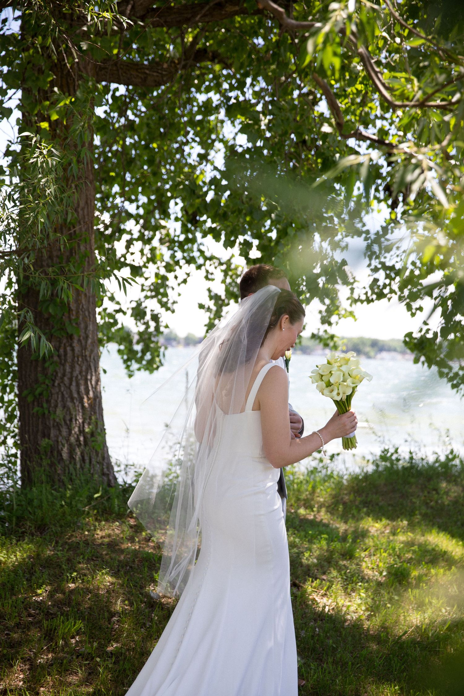 Bride in white gown and veil holds bouquet under green tree by waterside on her wedding day.