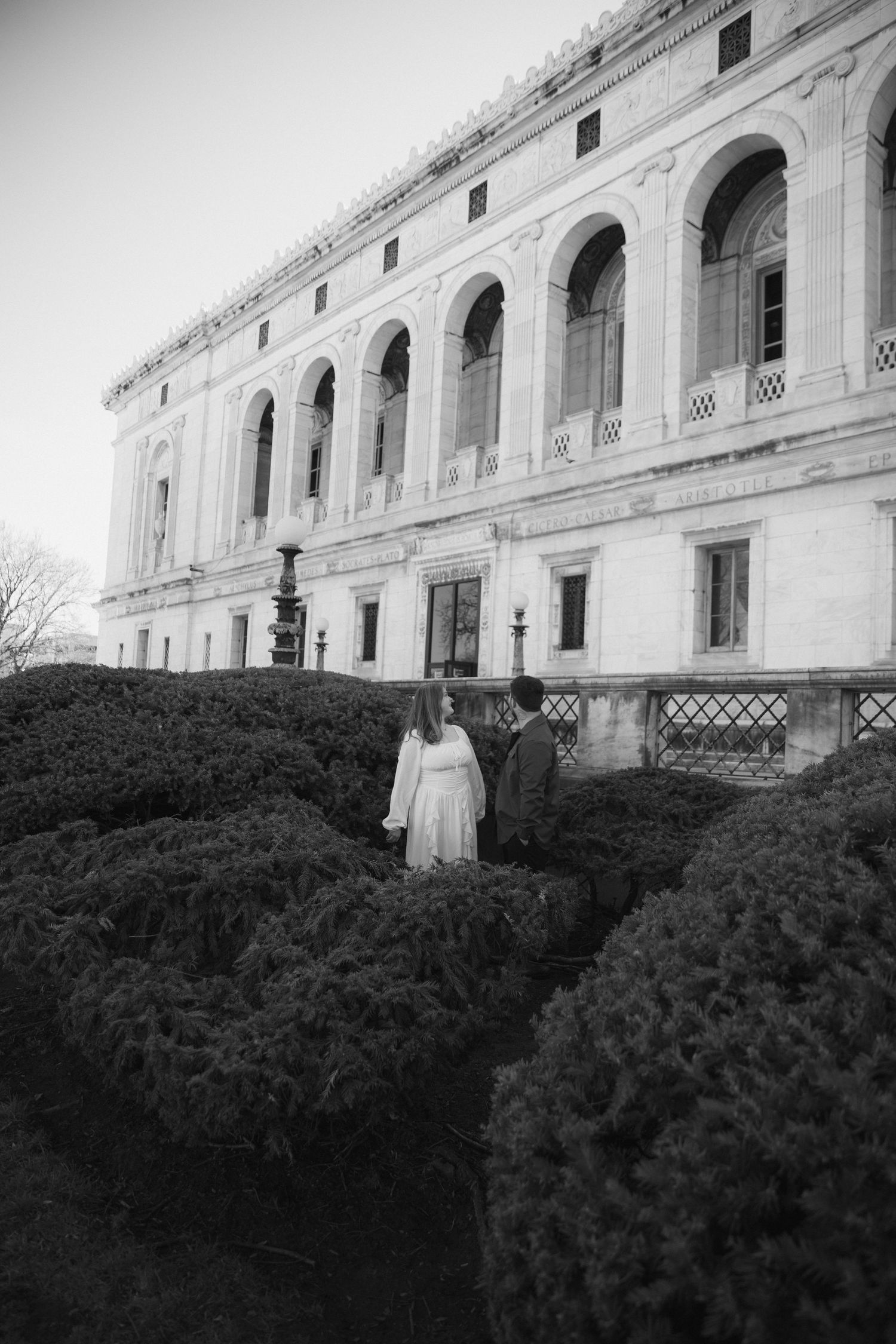 Black and white photo of a couple walking through manicured hedges before a grand neoclassical building.