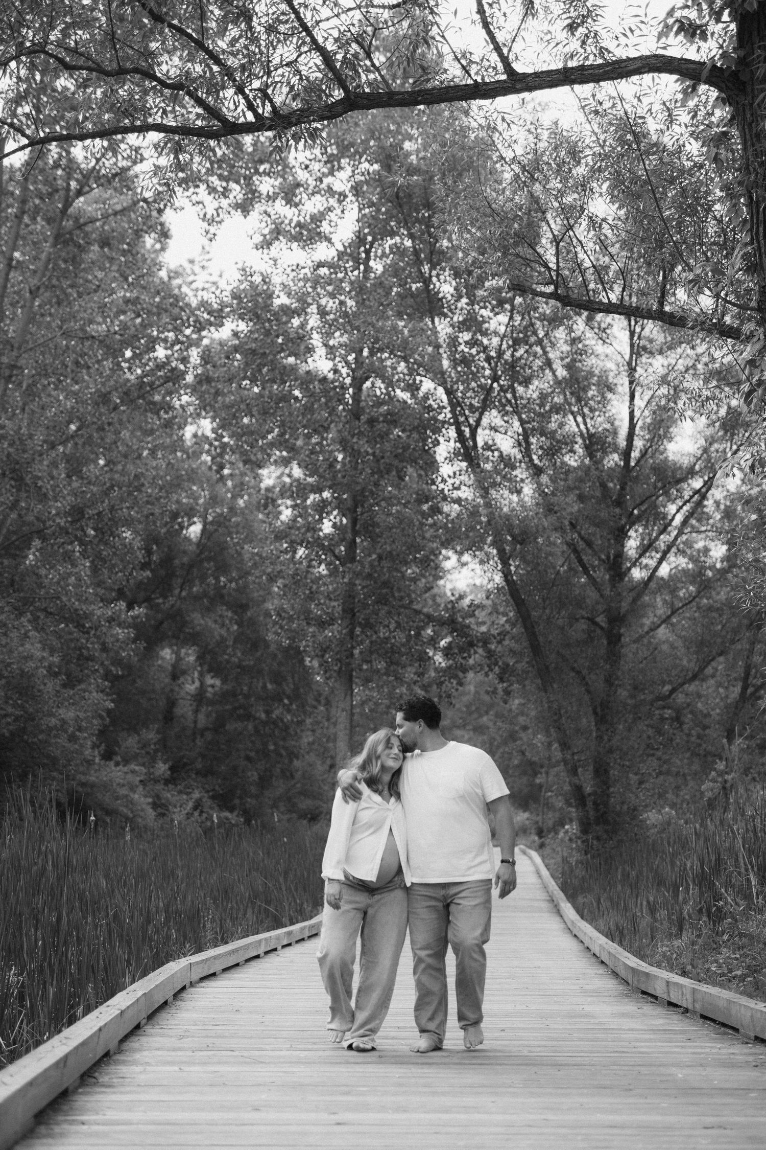 Black and white photo of a couple walking arm in arm on a wooden boardwalk path surrounded by lush trees.