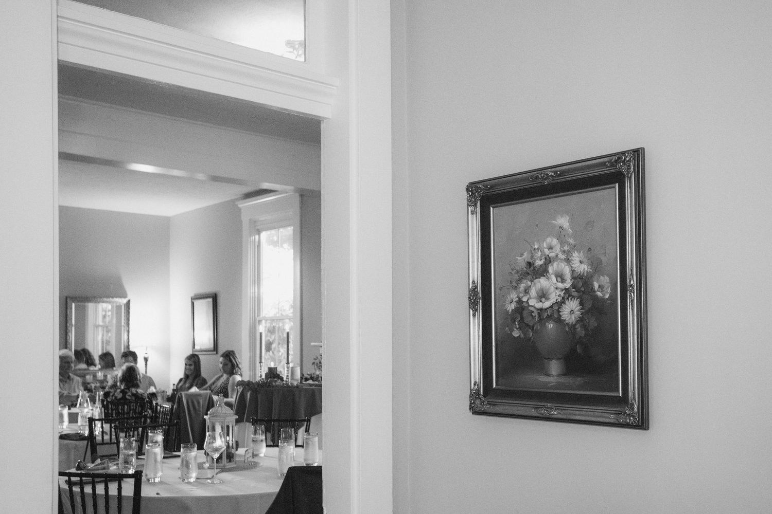 Black and white interior view of a hallway with framed artwork and a window showing an office space.