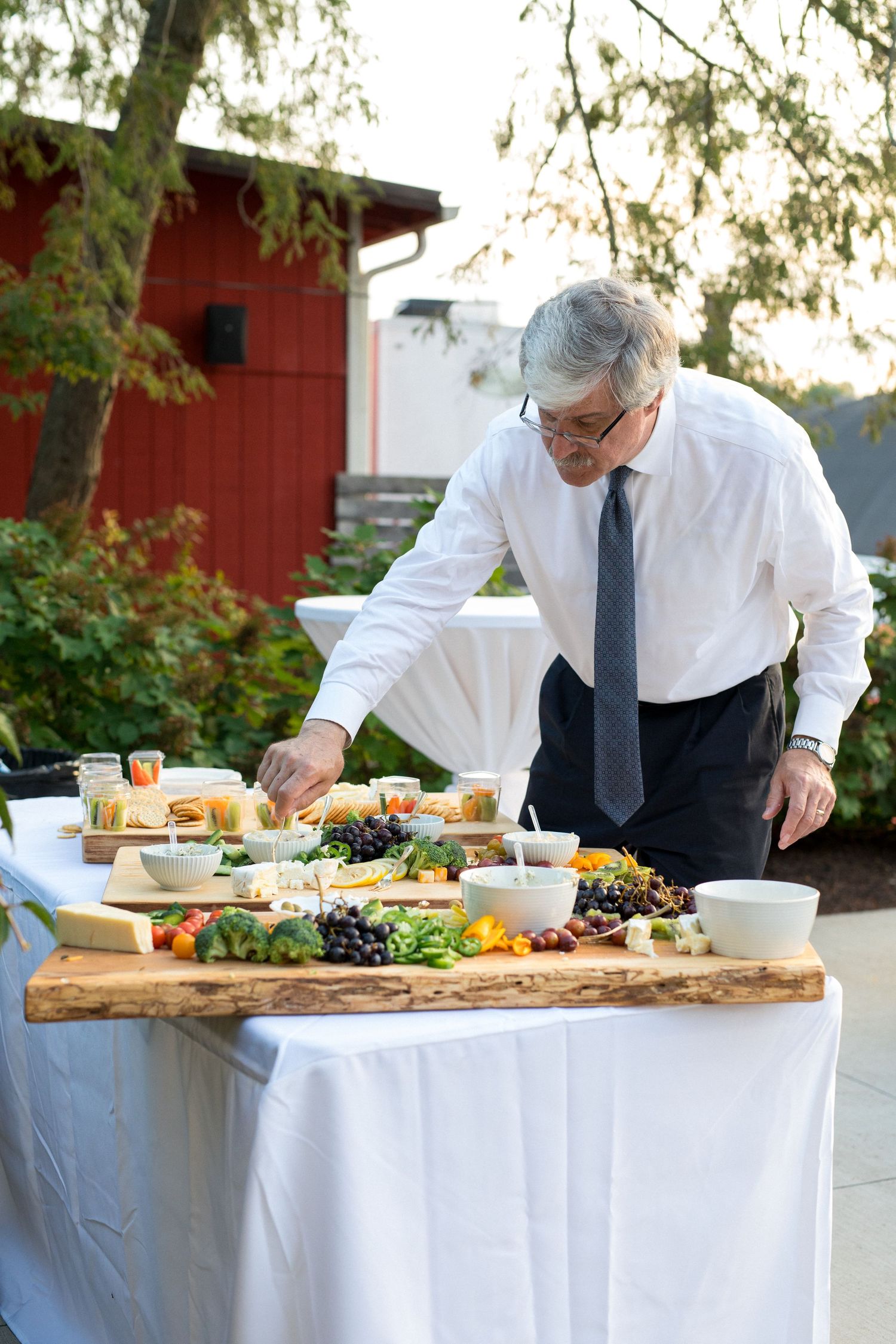 Professional chef arranges fresh ingredients and appetizers on white tablecloth at outdoor catering event.