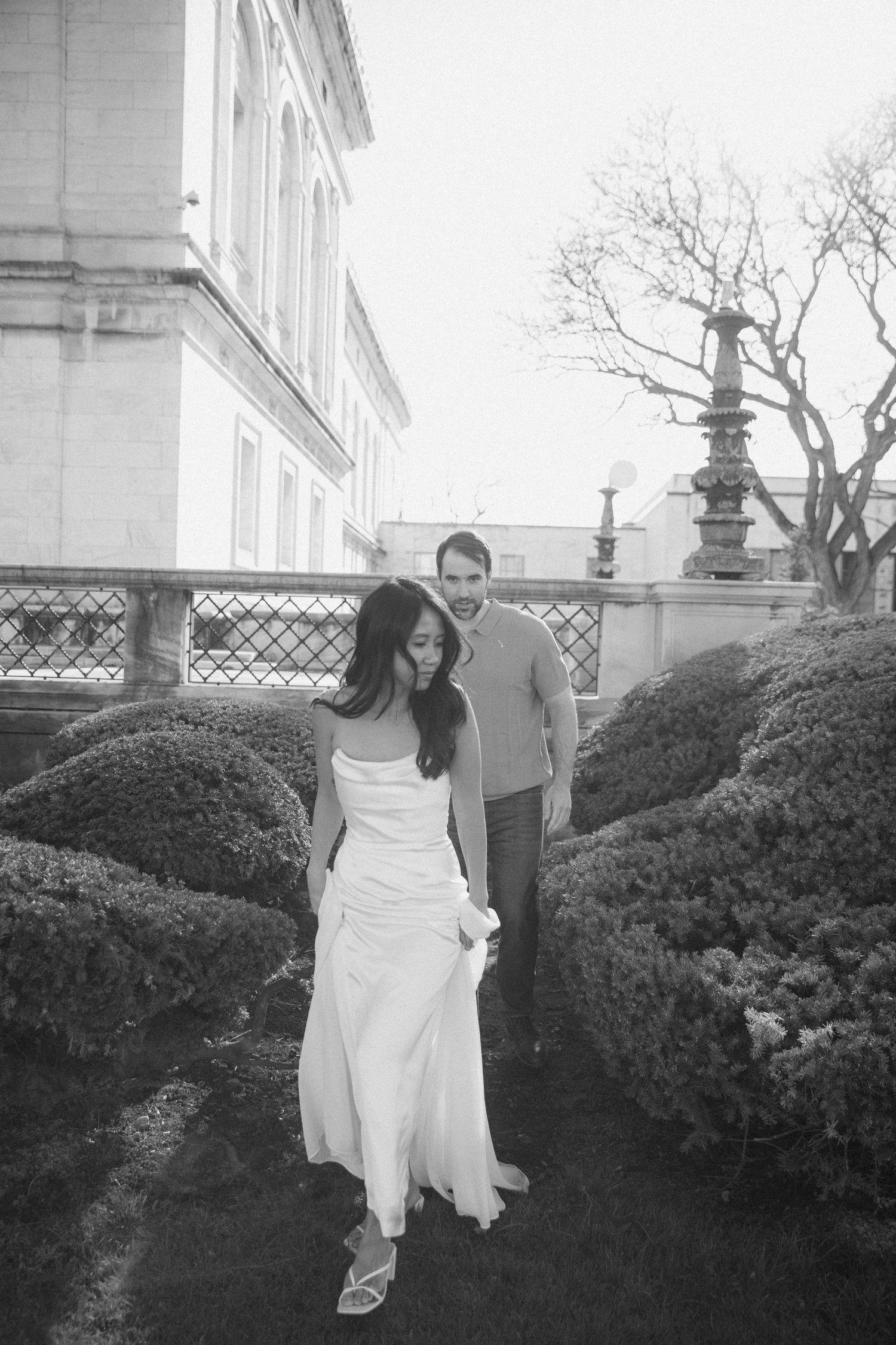 Bride in white gown walks through garden hedges as groom follows, elegant black and white photo.