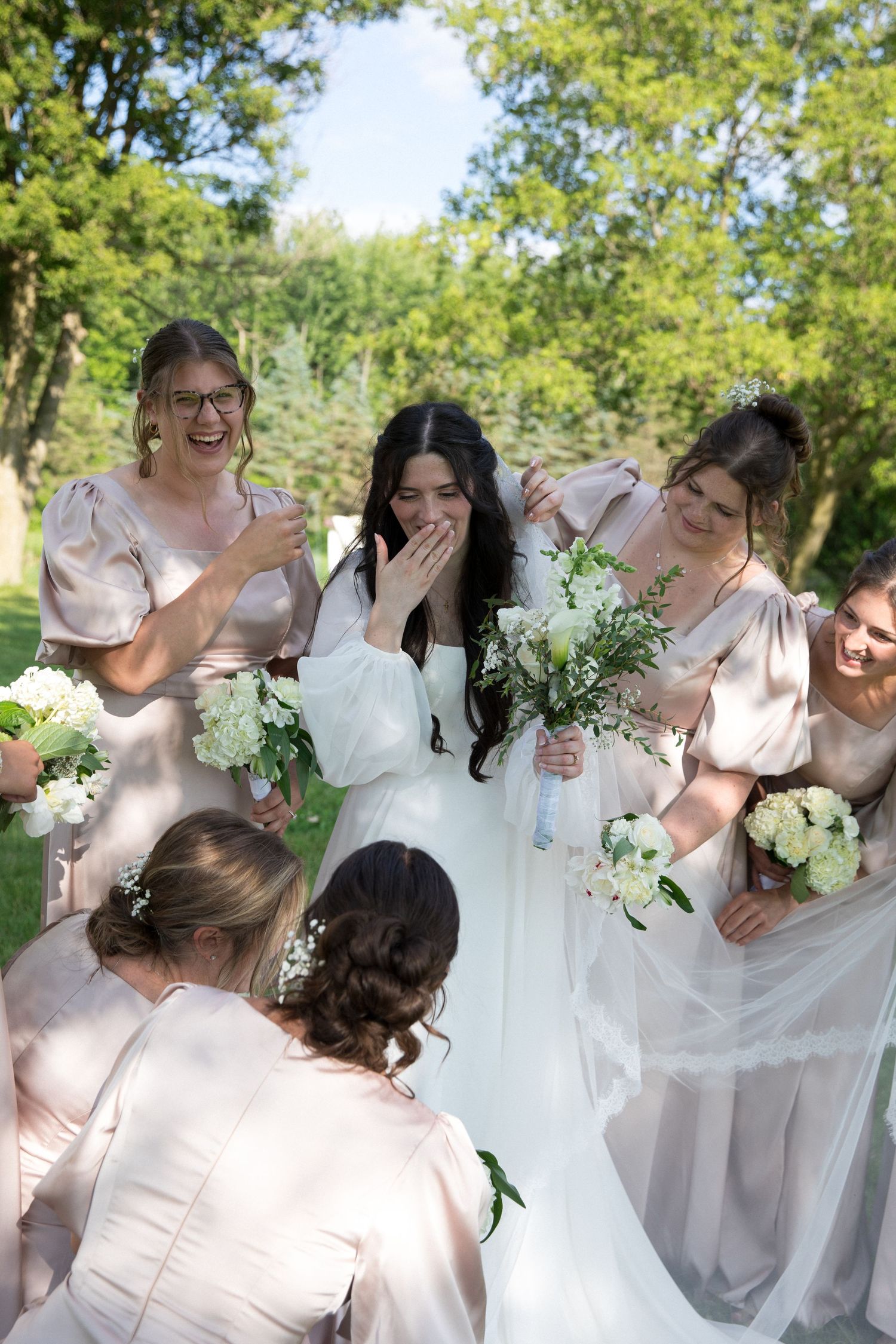 Bride in white gown laughing with bridesmaids in blush dresses holding white bouquets outdoors.