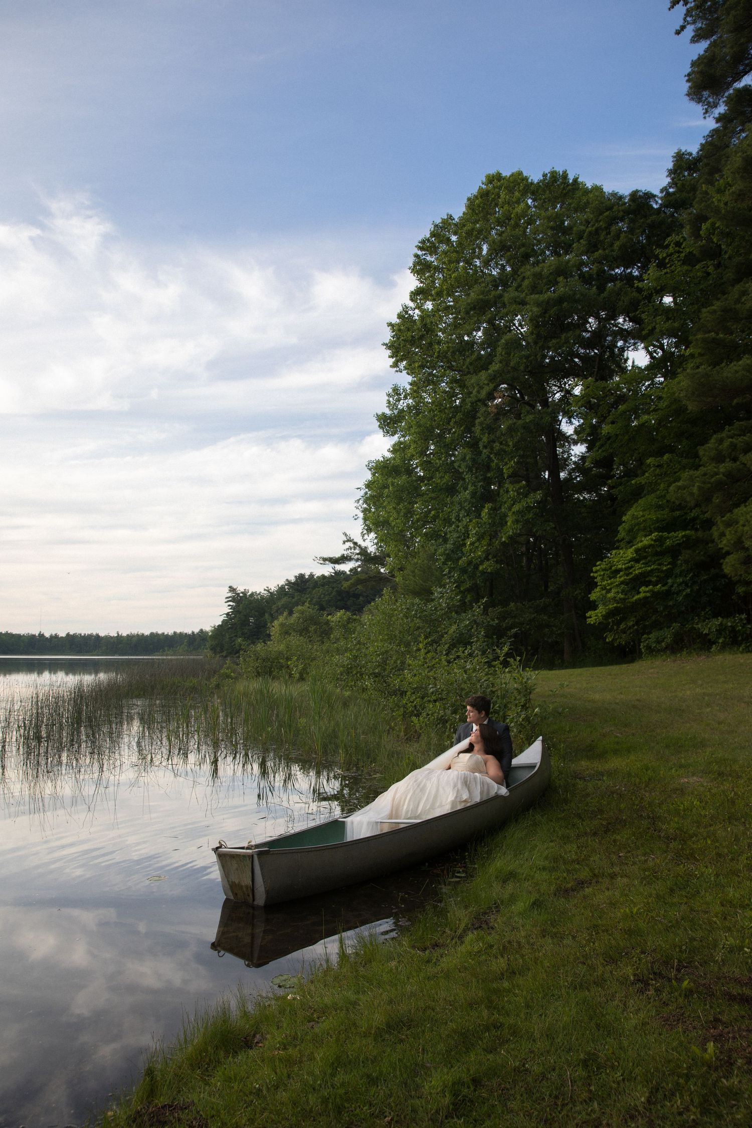 Bride in white wedding dress sitting in a canoe by a calm lake shore surrounded by lush green trees at dusk.