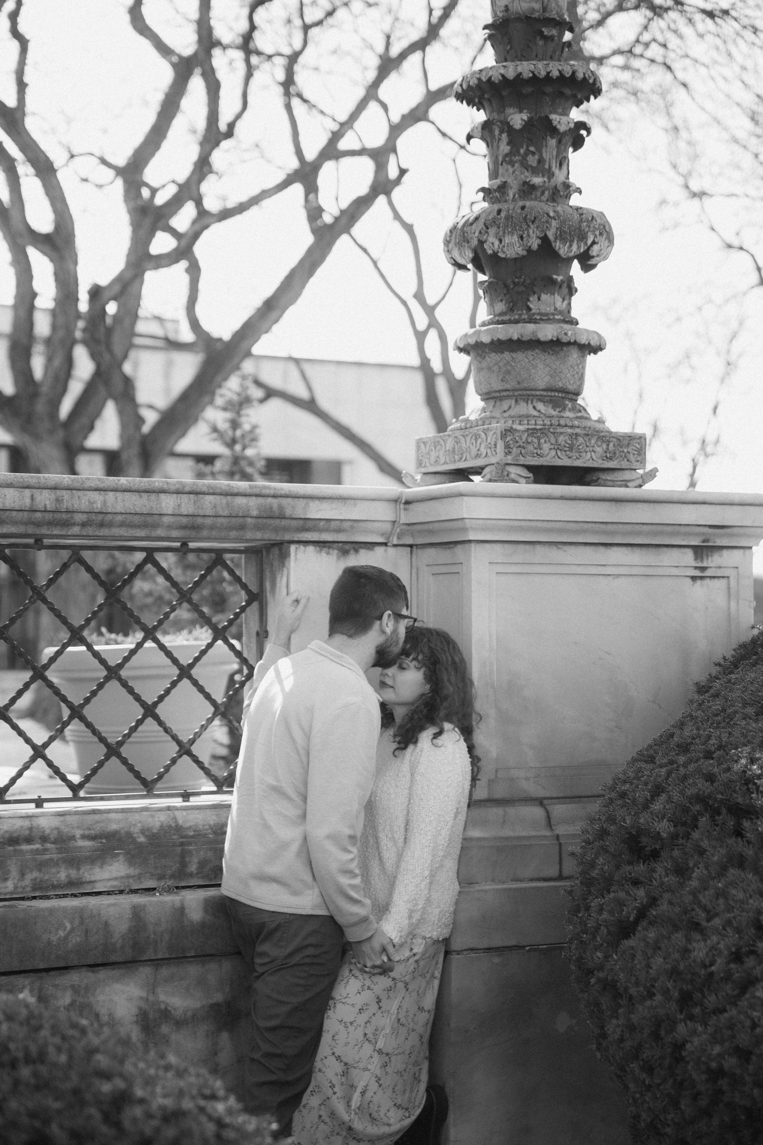 Black and white photo of a couple sharing a romantic moment near an ornate stone pillar and decorative fence outdoors.
