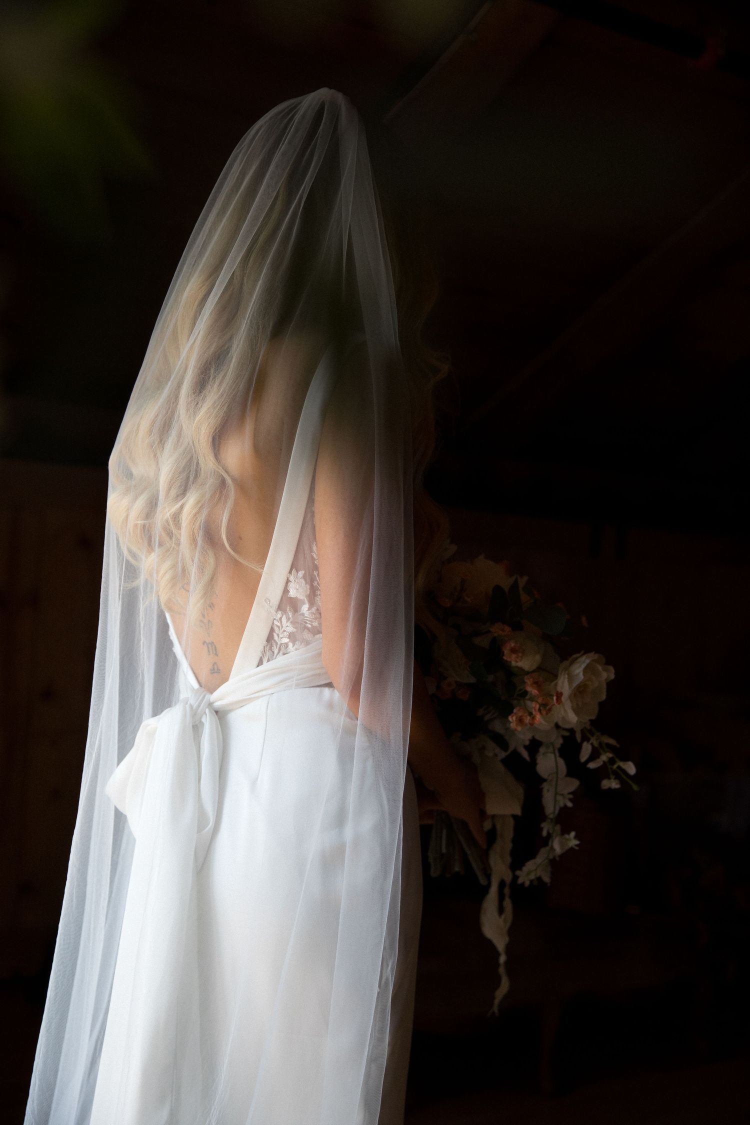 A bride in a white wedding dress and flowing veil stands in dramatic lighting creating an ethereal silhouette.