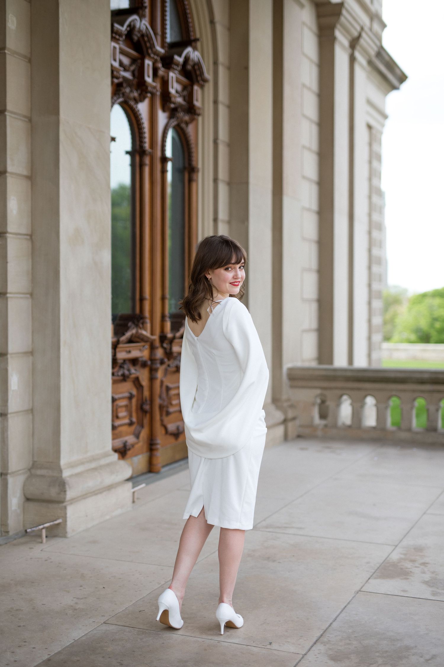 Someone in a white outfit poses in a dramatic sequence along an elegant colonnade with stone archways and columns.