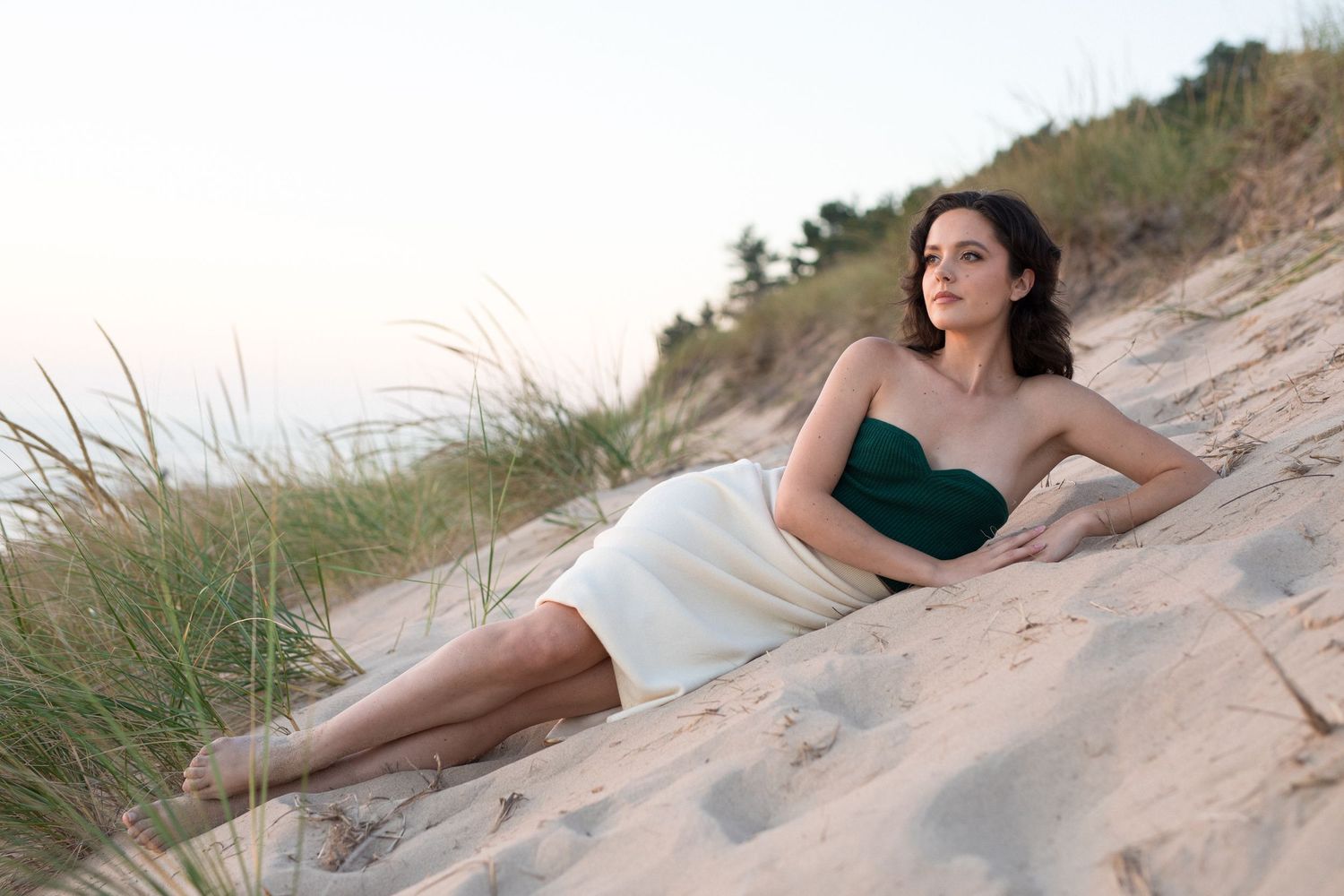 A person in a green dress poses elegantly on sandy dunes with beach grass at sunset.