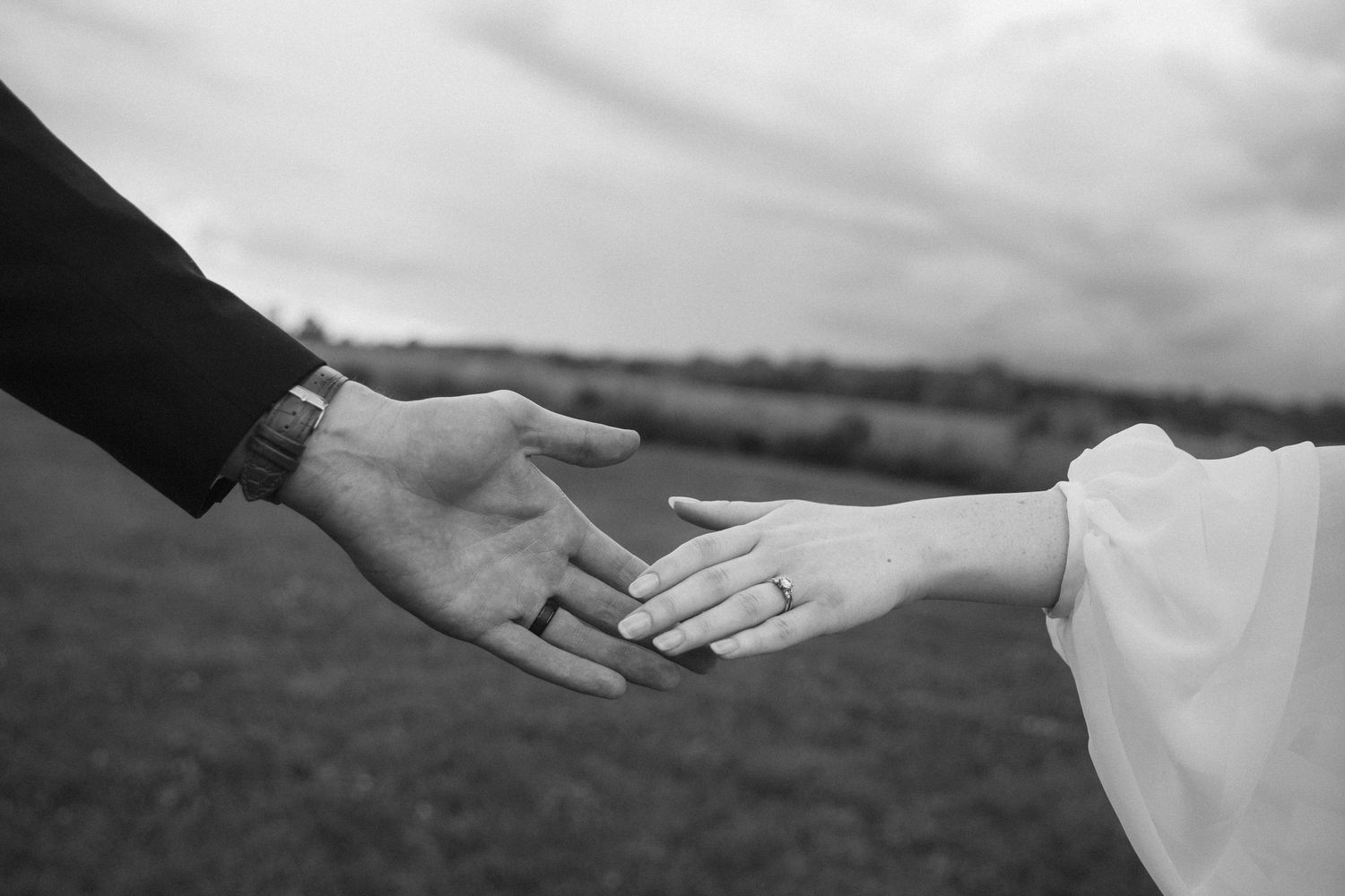 Black and white photo of a bride and groom holding hands outdoors, showcasing a wedding ring on the brides finger.