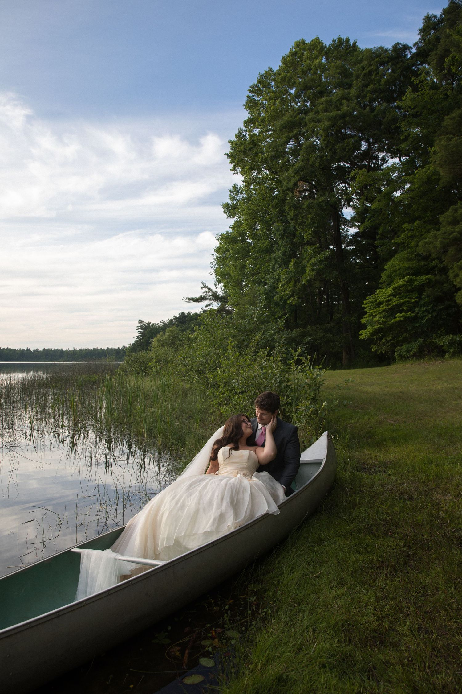 Bride and groom share a romantic kiss in a canoe by a serene lake surrounded by lush green trees at dusk.