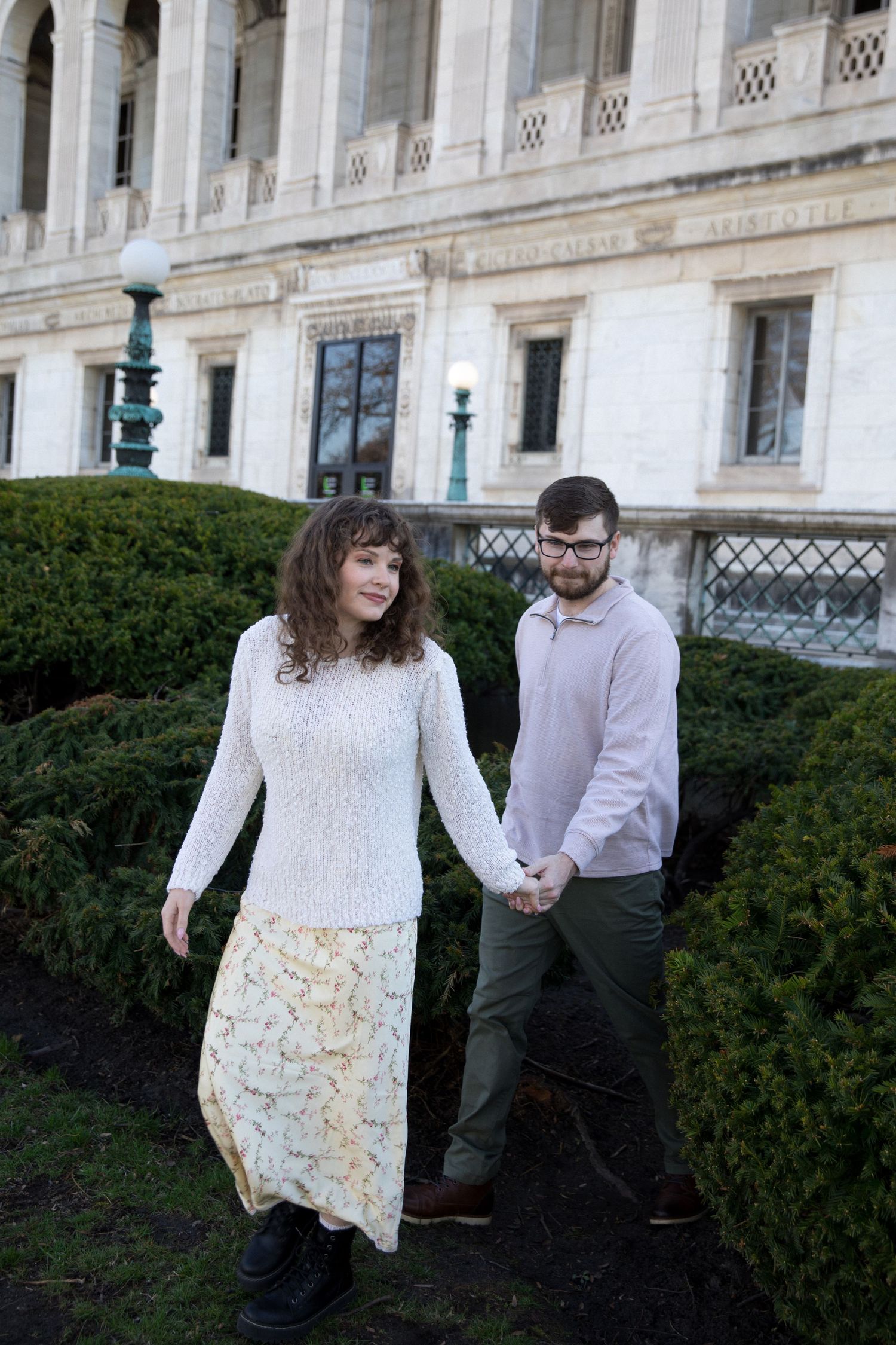 A couple holds hands walking through a garden path beside a grand historic building, woman in white lace dress and man in casual attire.
