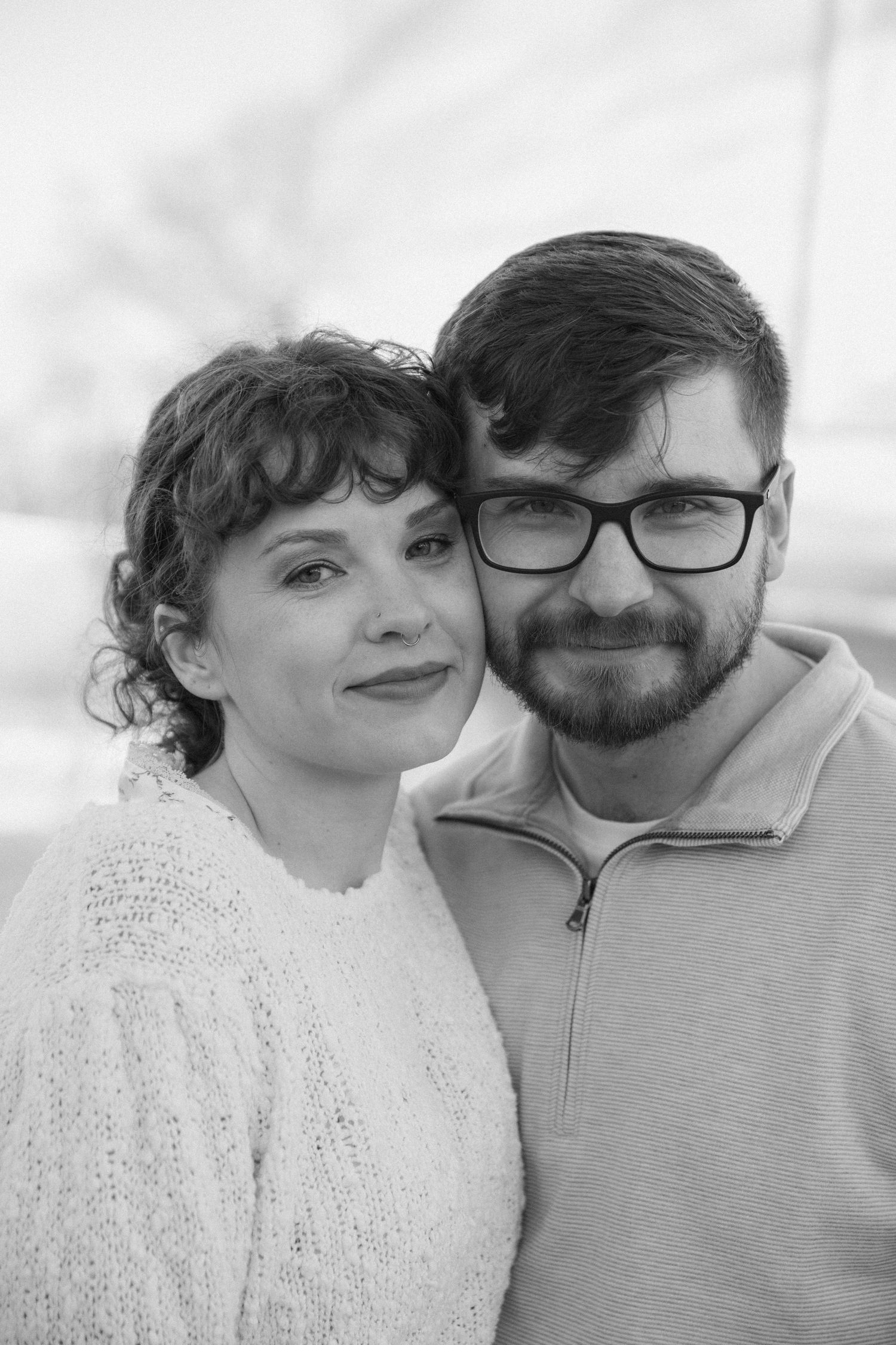 Black and white portrait of a smiling couple, woman with curly hair and man with glasses and beard posing together.
