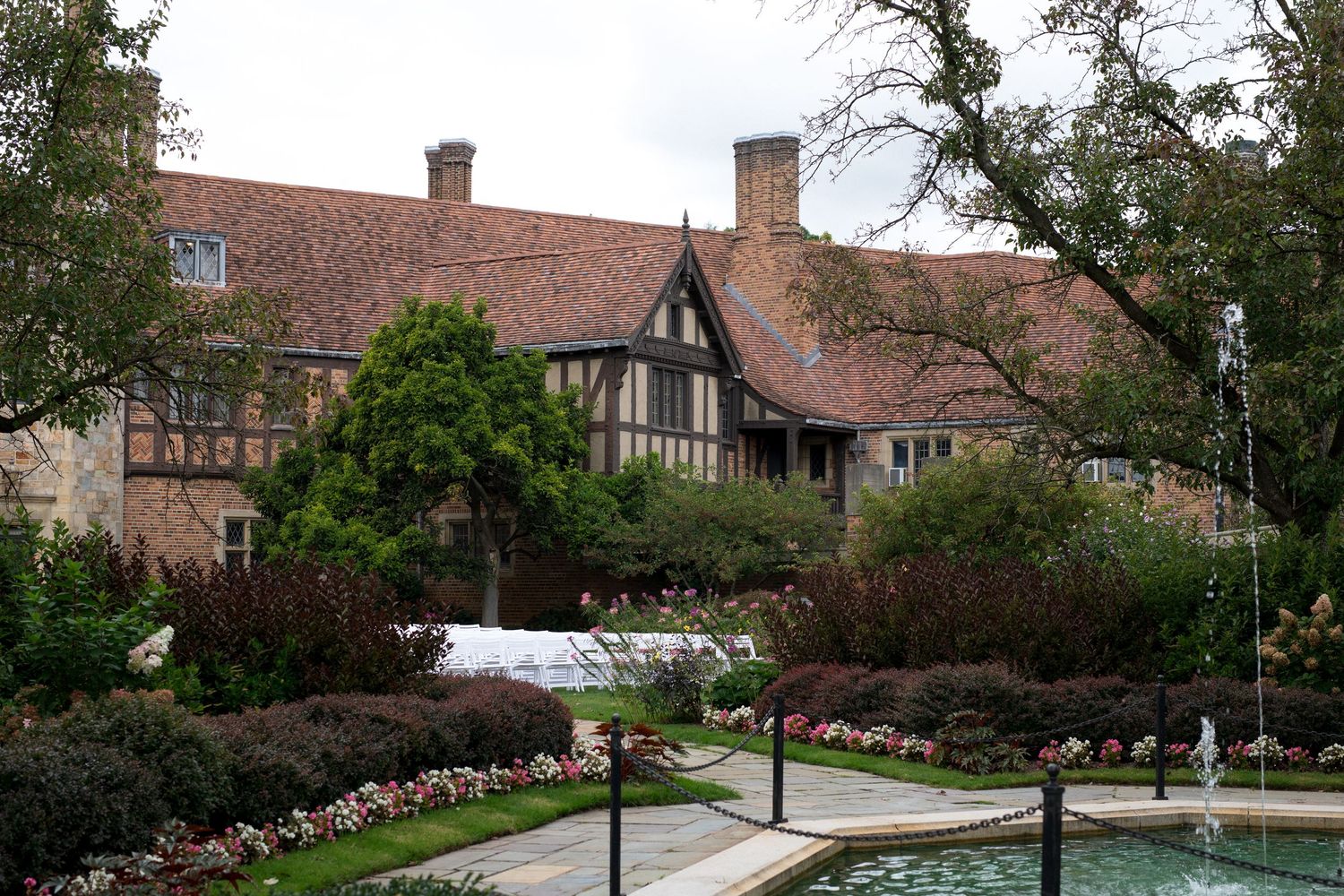 Tudor-style mansion with red brick and timber framing surrounded by manicured gardens and a decorative pool.