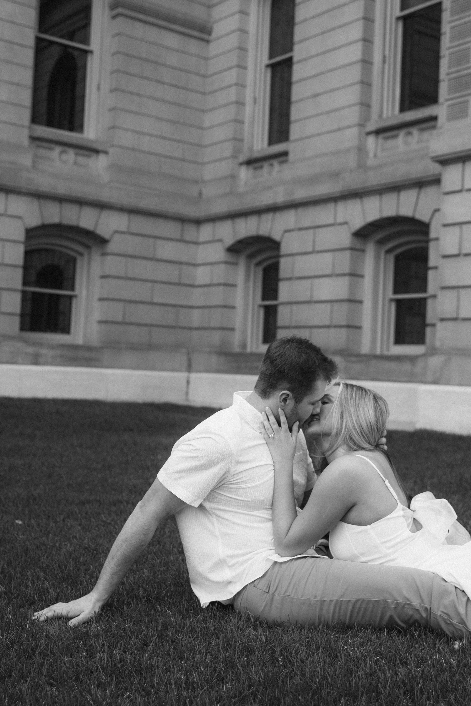 A romantic couple shares a kiss while sitting on grass in front of a classic stone building in black and white.