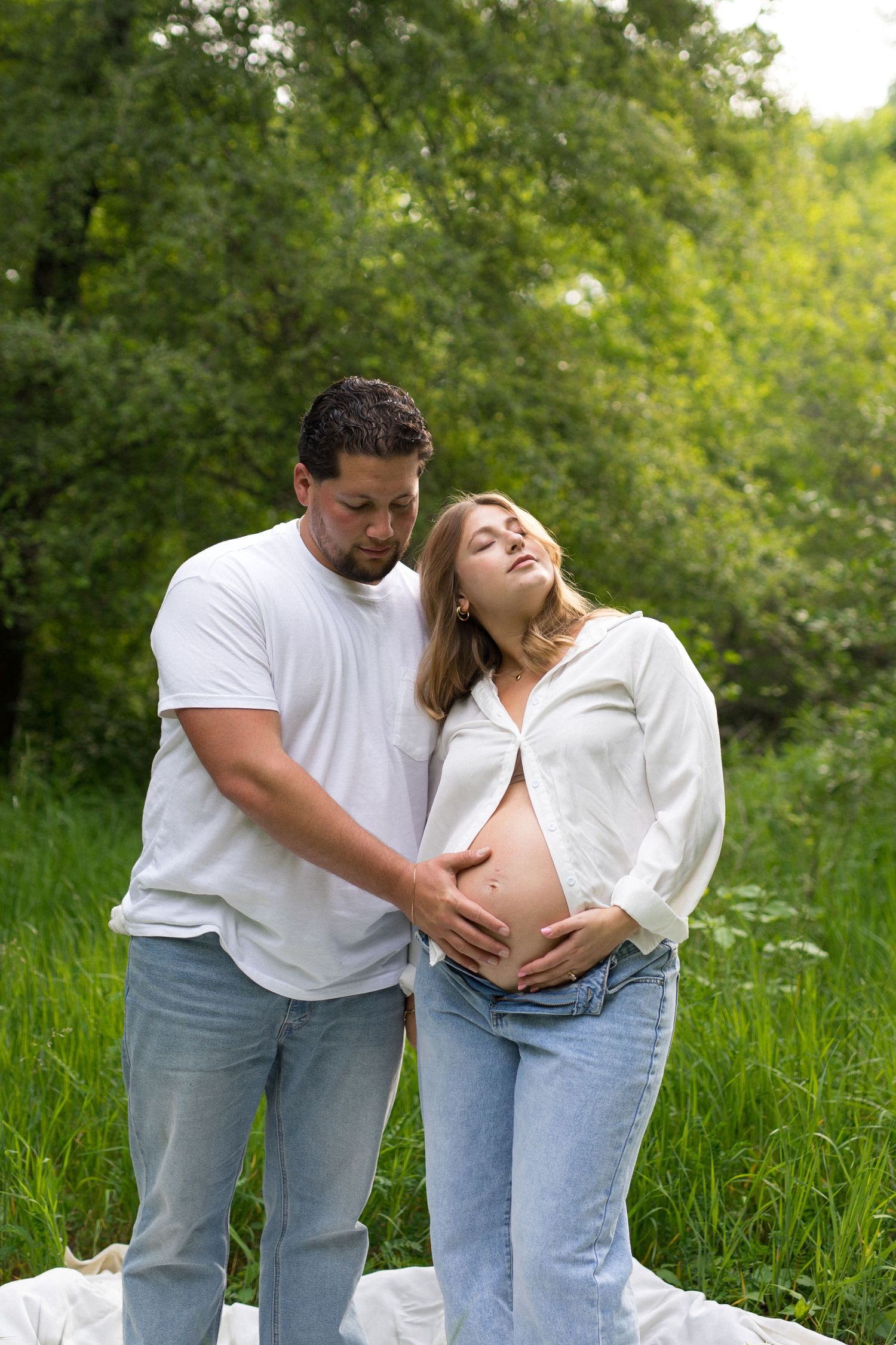 A couple tenderly cradles a pregnant belly outdoors, surrounded by lush green trees during a maternity photo shoot.