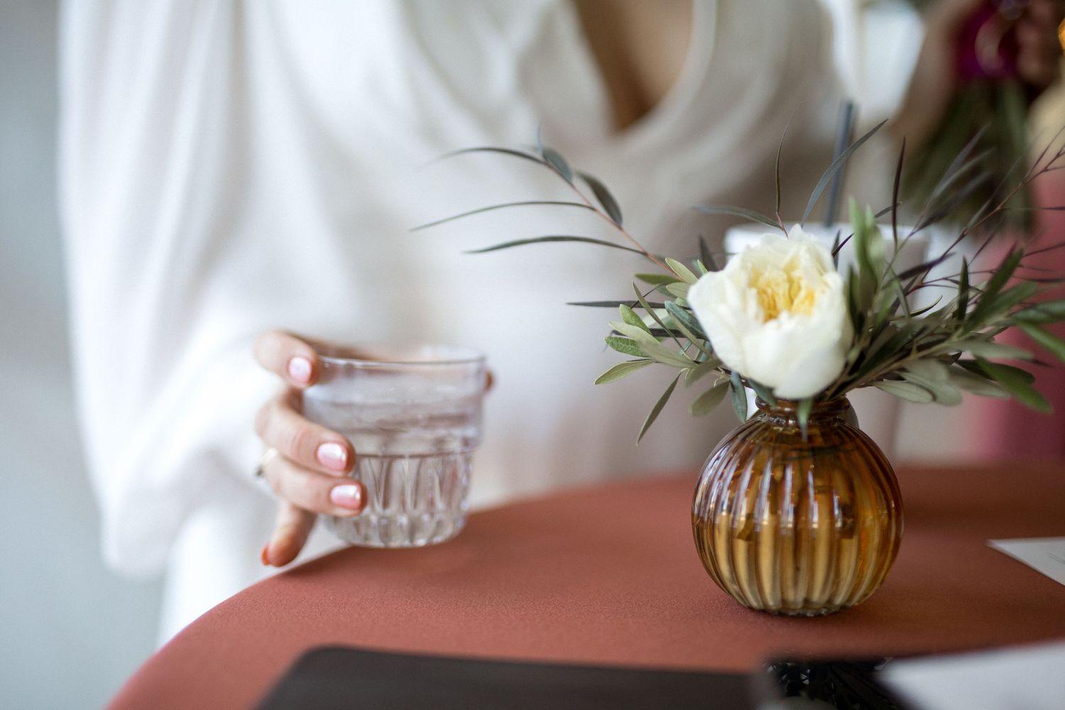 A hand holds a delicate white teacup against a soft, neutral background with a floral arrangement in view.