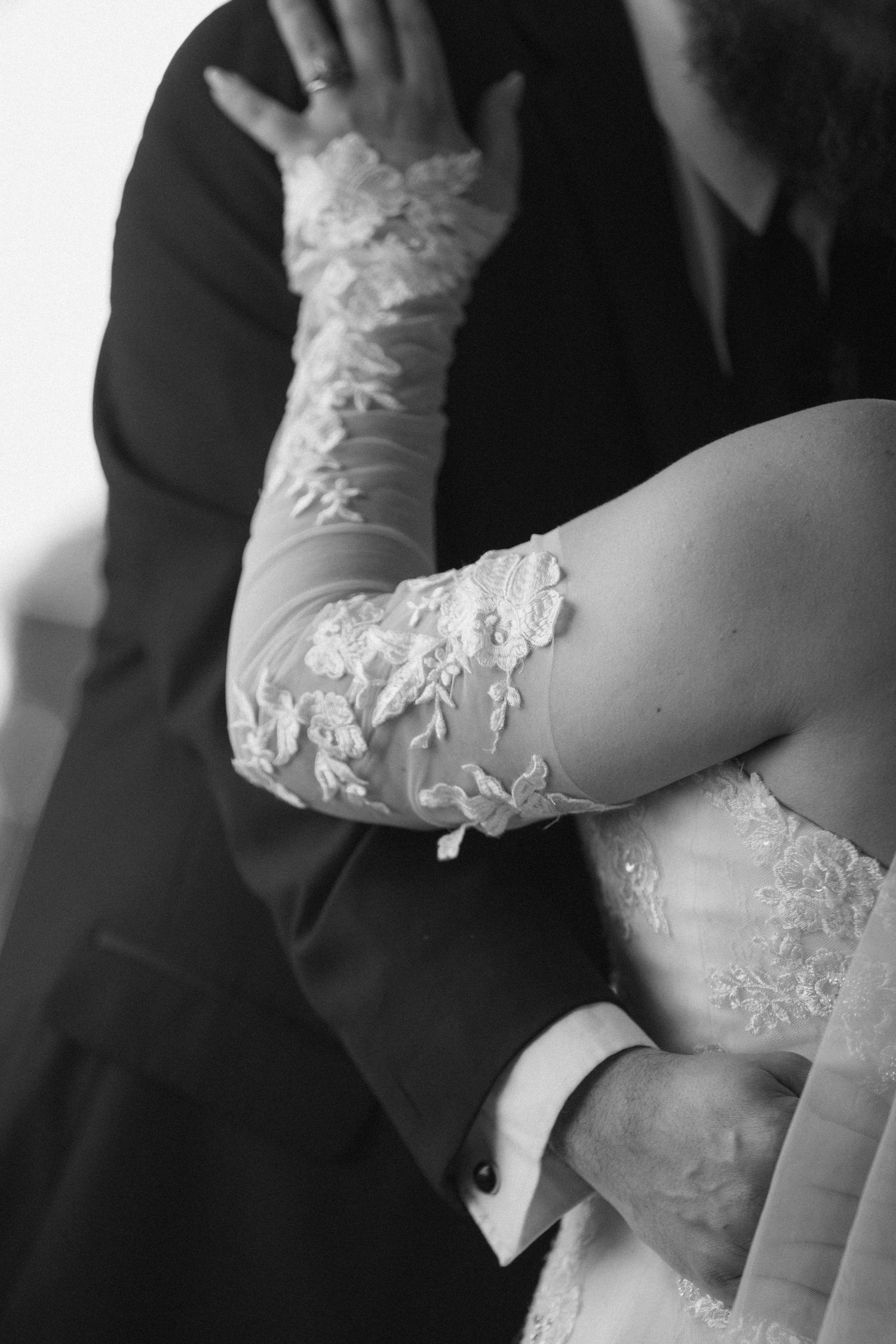 Black and white photo of bride in lace gloves embracing groom in suit at wedding ceremony.