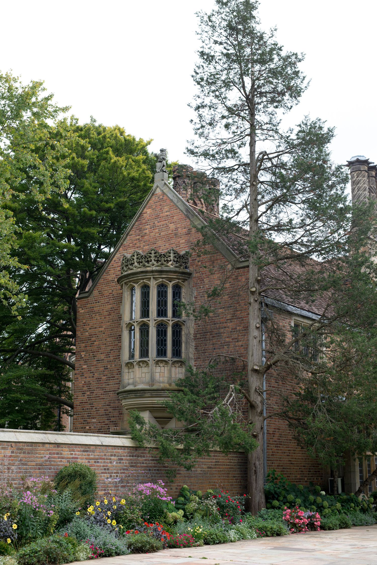 Historic brick mansion with Gothic windows surrounded by colorful flower gardens and tall trees.
