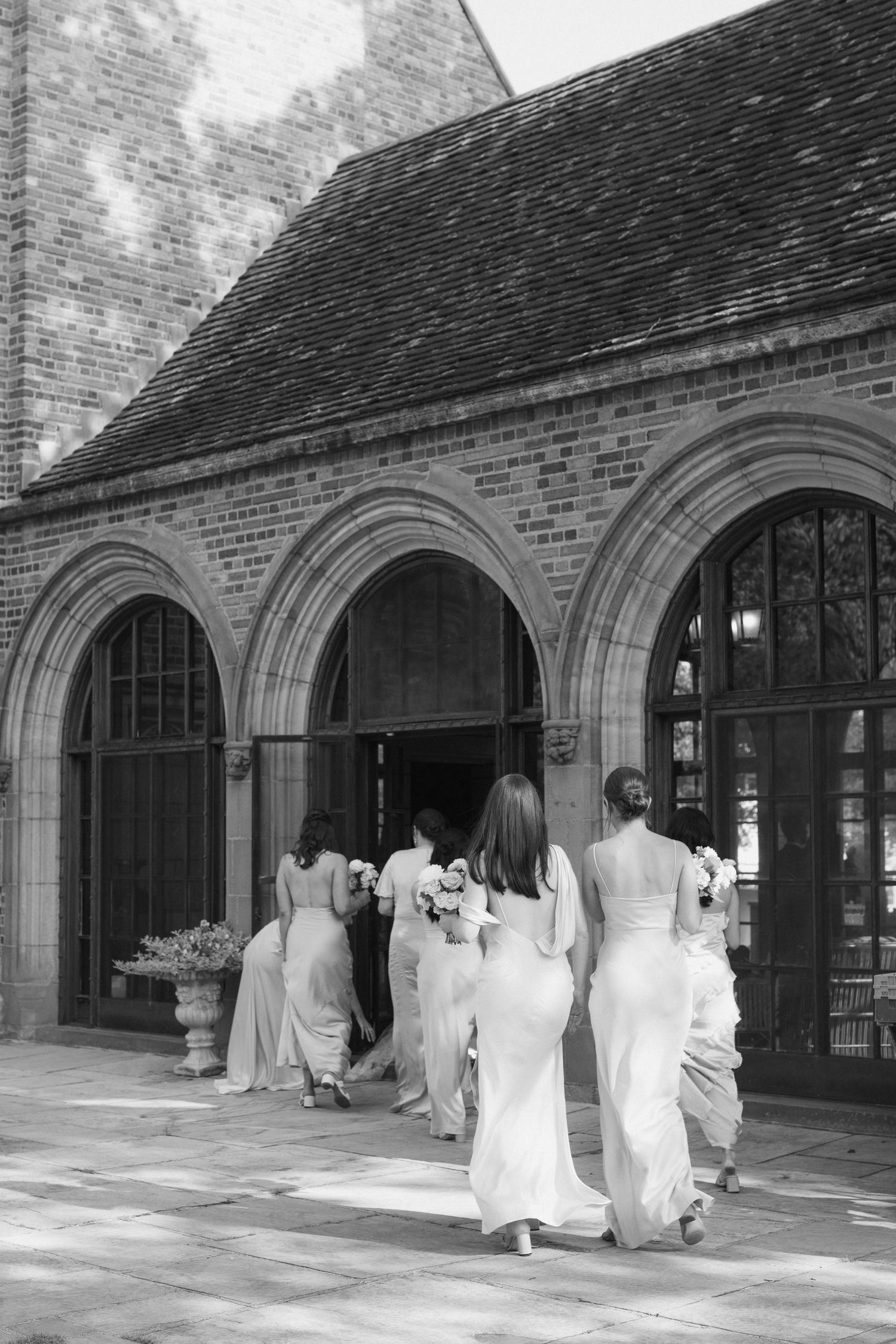 Wedding party members in white dresses gather under brick archways of a historic building.