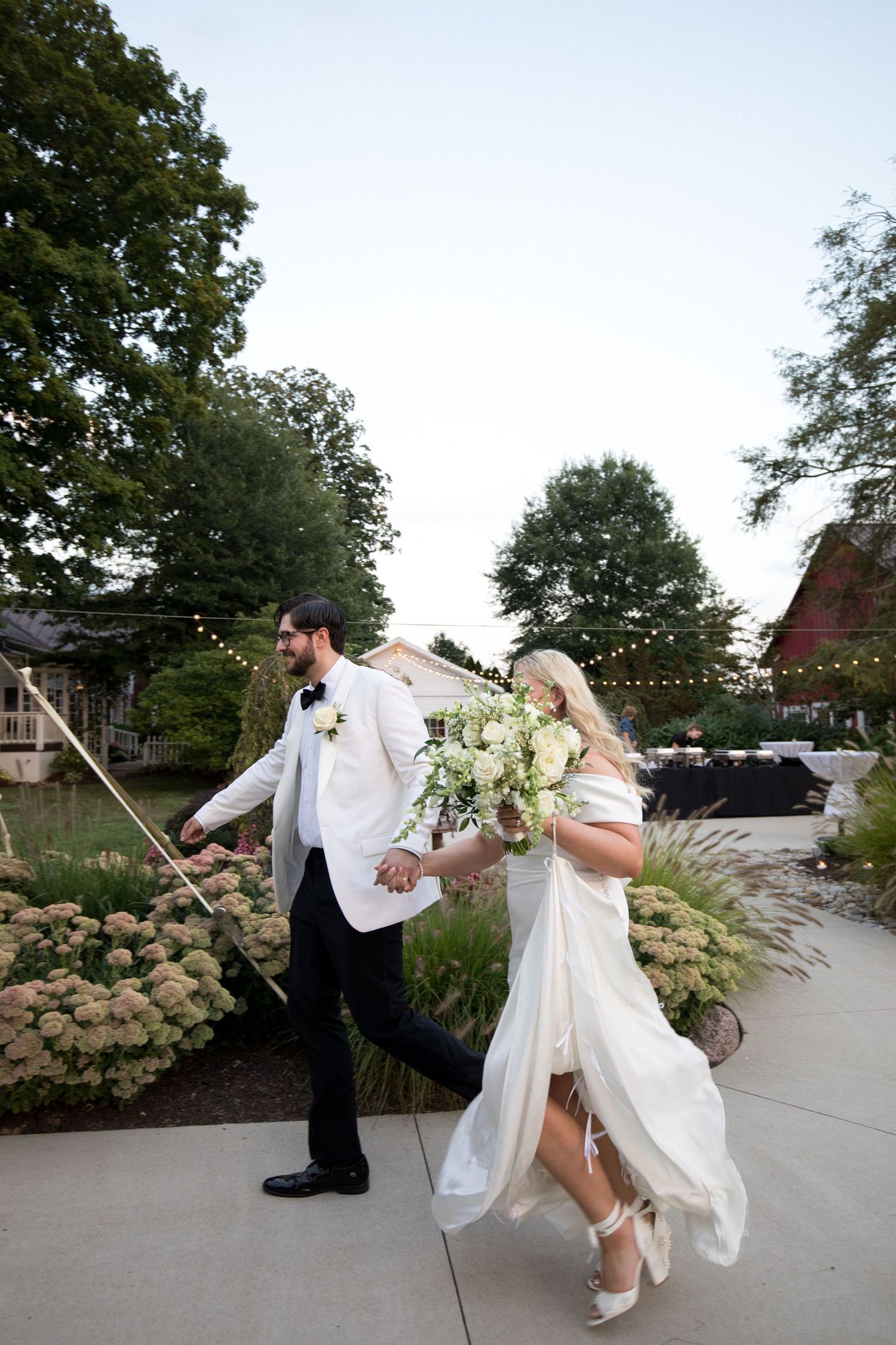 A romantic wedding couple dances together on an outdoor patio surrounded by hydrangea bushes and string lights at dusk.