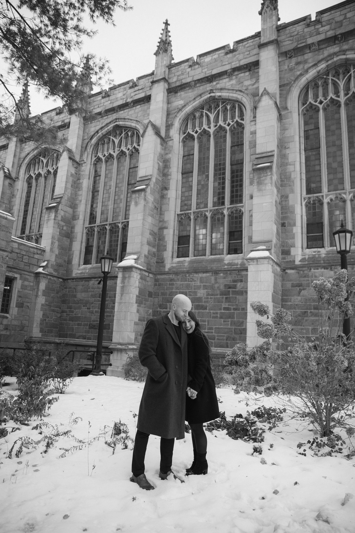 Two figures walk along a snowy path beside a Gothic stone building with arched windows and architectural details.
