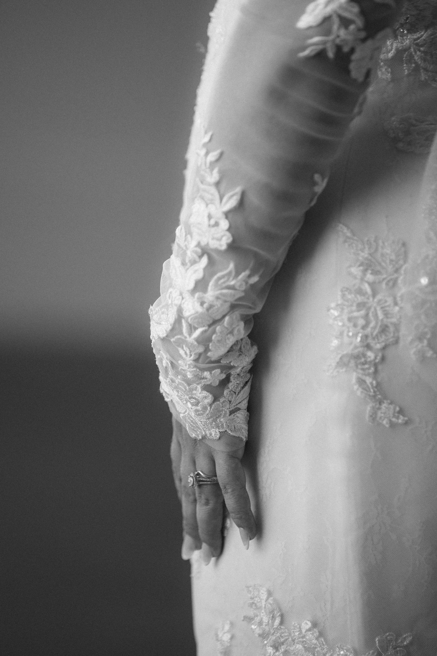 Close-up black and white photo of a bride's lace wedding dress sleeve and gloved hand with engagement ring.