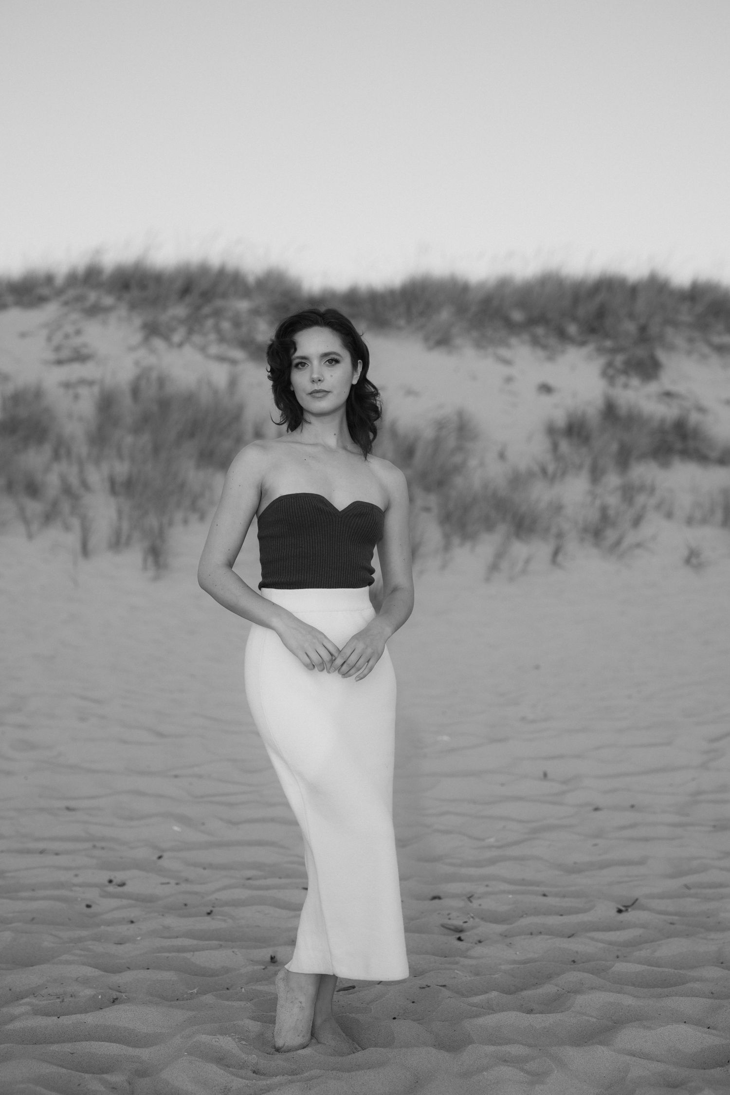 Woman in black top and white skirt poses elegantly on sandy beach dunes in black and white photography.