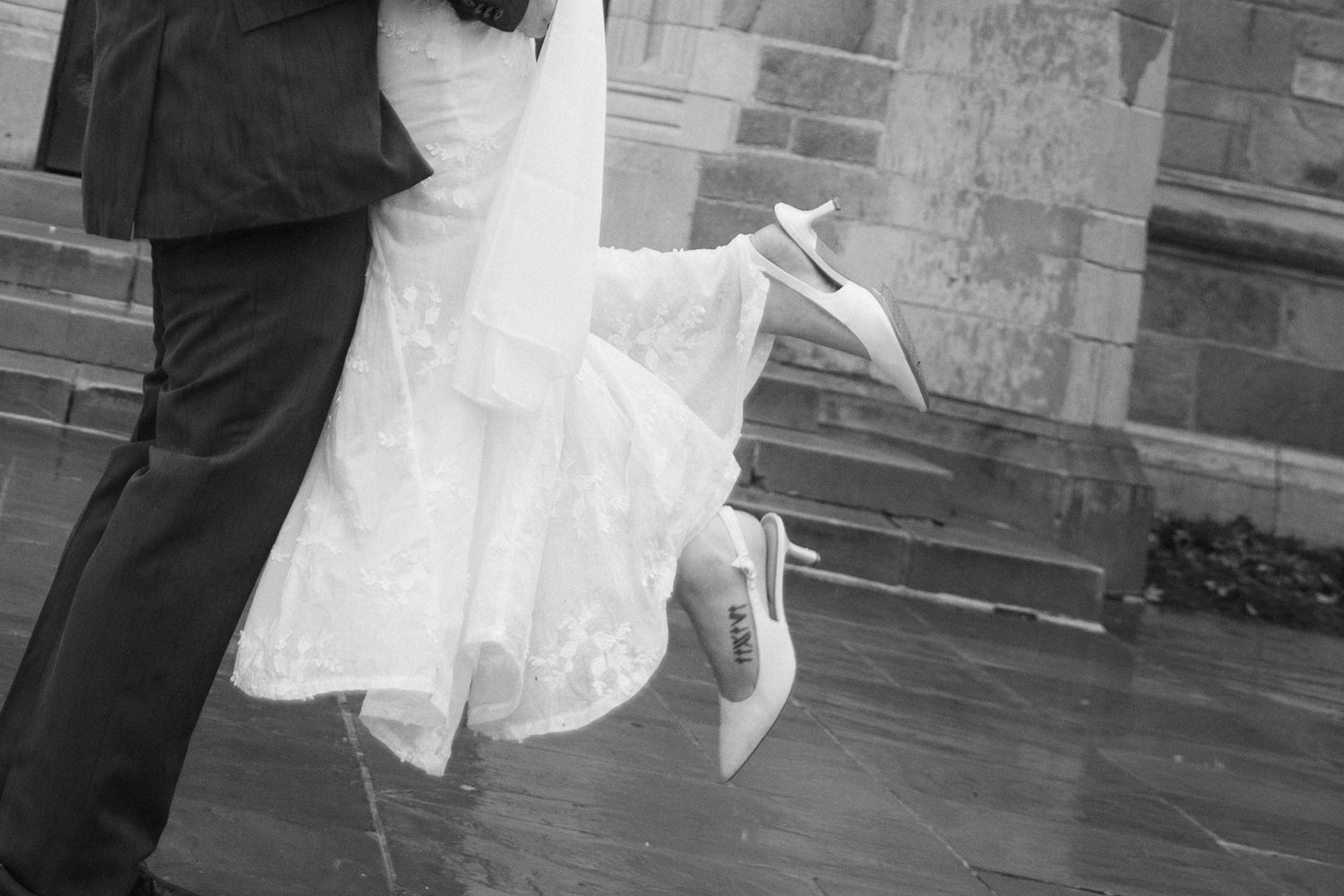 Black and white photo of a bride's flowing white dress and groom's dark suit as they embrace on stone church steps.
