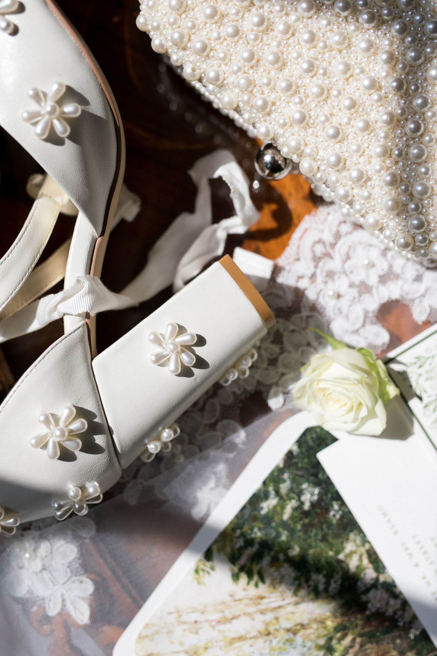 White wedding shoes with delicate floral details rest among scattered pearls and greenery on a wooden surface.