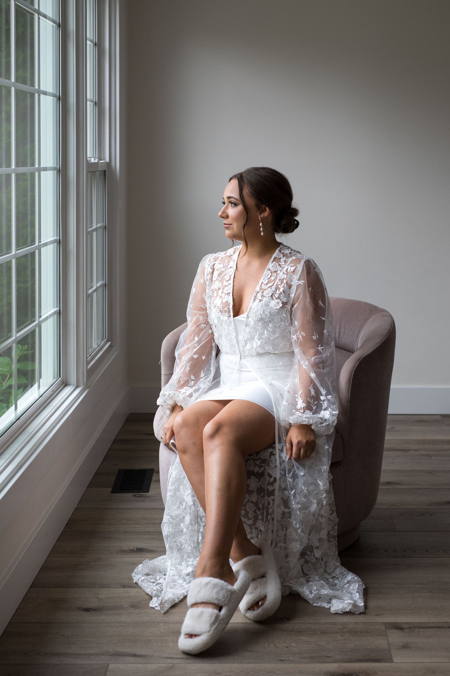 A person in a white lace robe sits by a window wearing fuzzy slippers in an elegant boudoir setting.