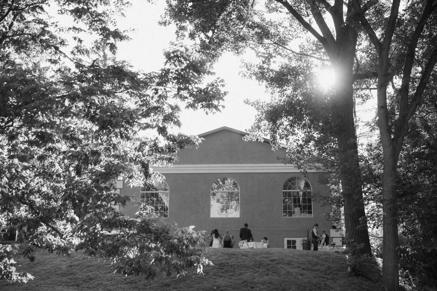 Black and white photograph of a house with sunlight streaming through trees in a serene backyard setting.