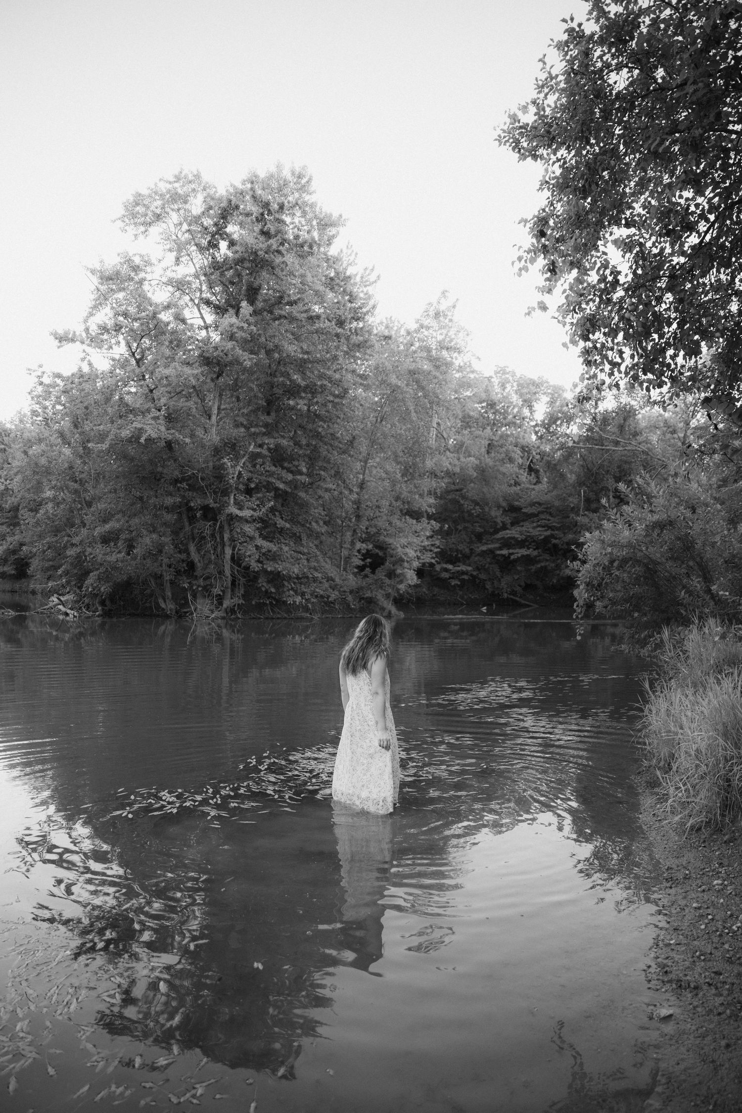 A haunting black and white photograph shows a white dress standing in still water surrounded by trees.