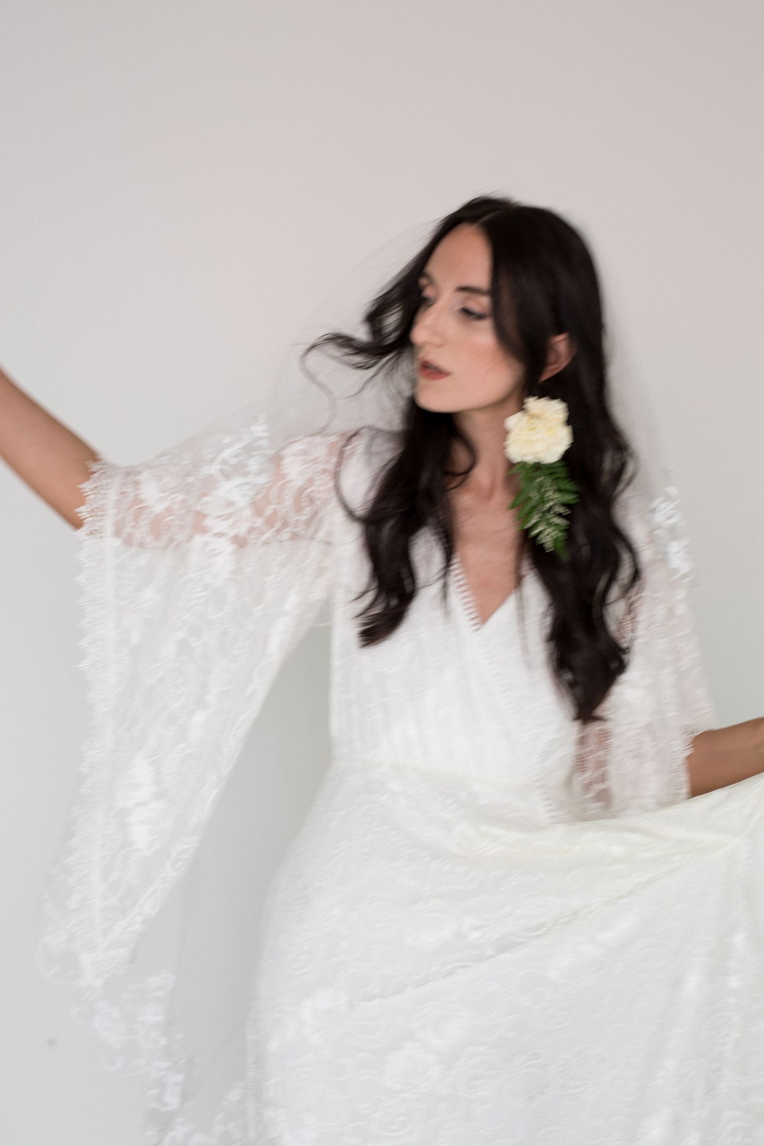 Bride in white lace kimono-sleeve gown with flower hair accessory poses gracefully against light background.