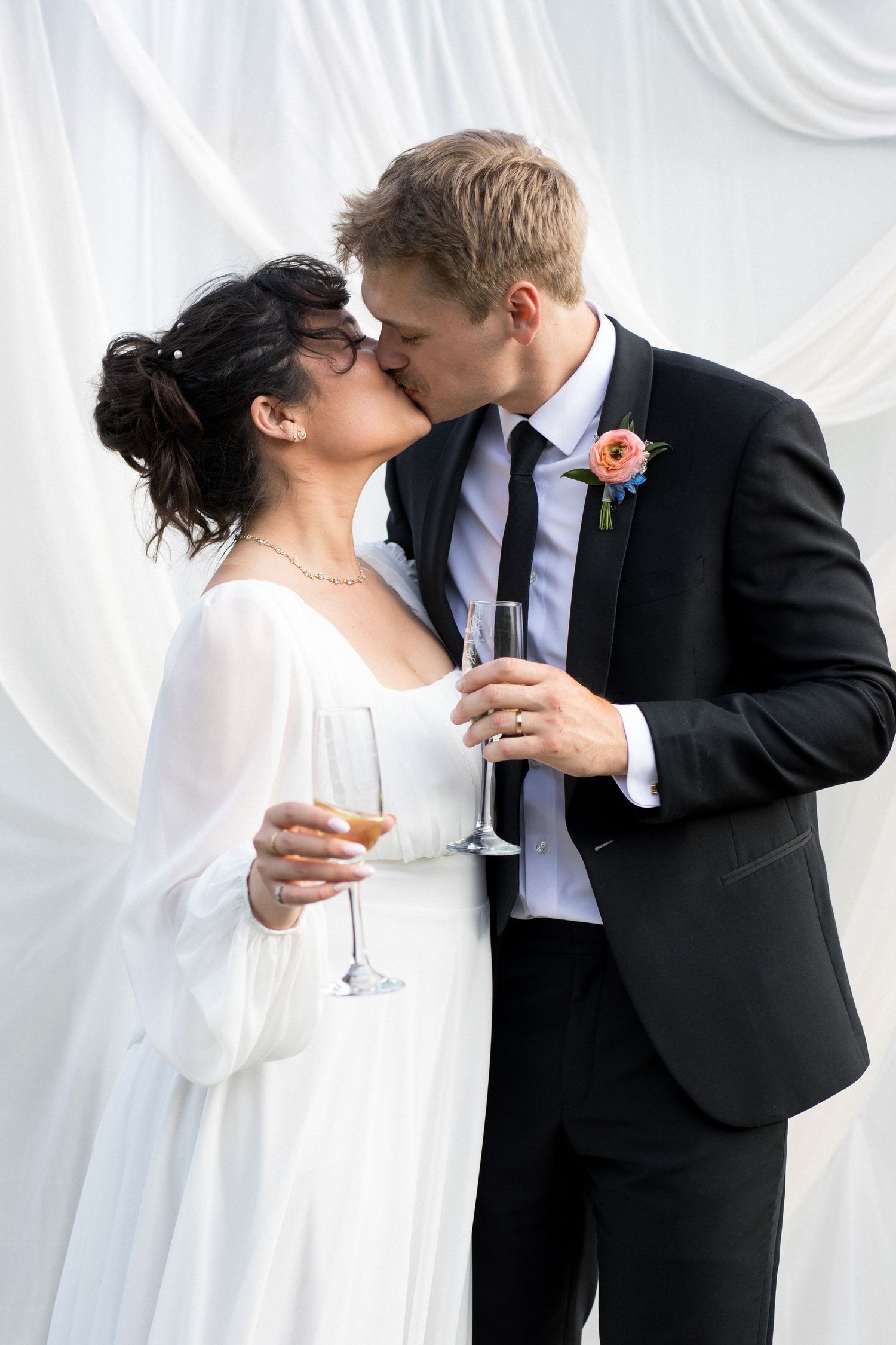 A bride and groom embrace and kiss on their wedding day, with the bride wearing a white dress and the groom wearing a black suit.