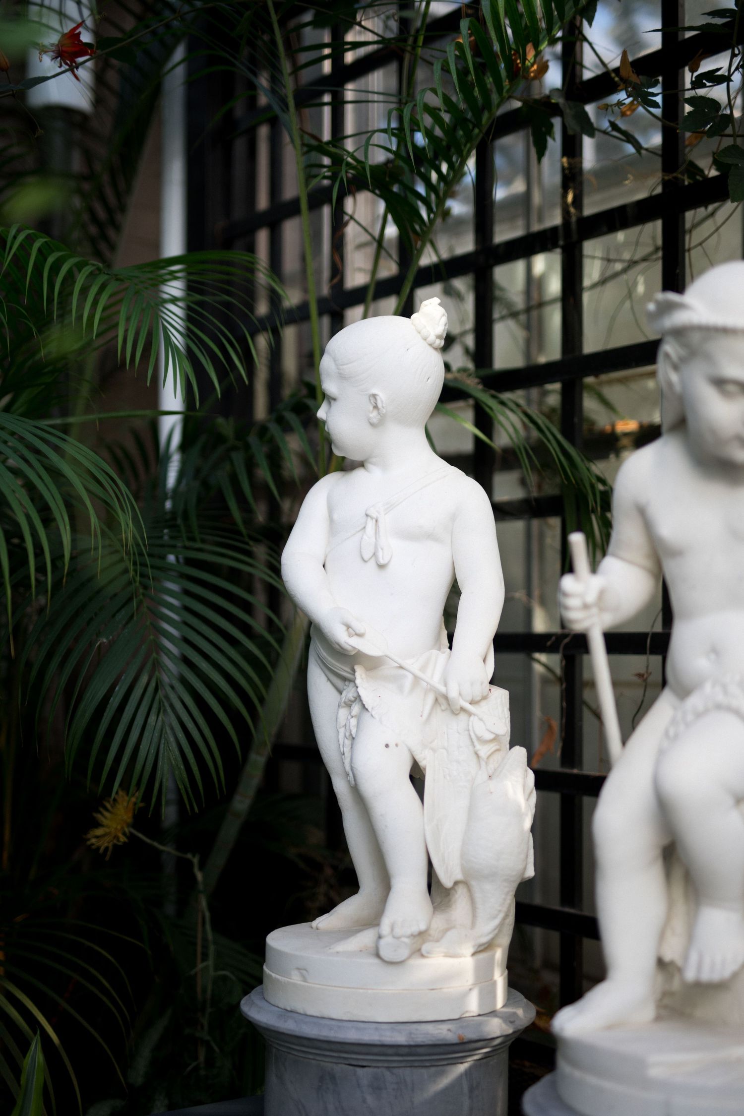 White marble statues of cherubs and figures displayed among tropical palm plants in a garden setting.