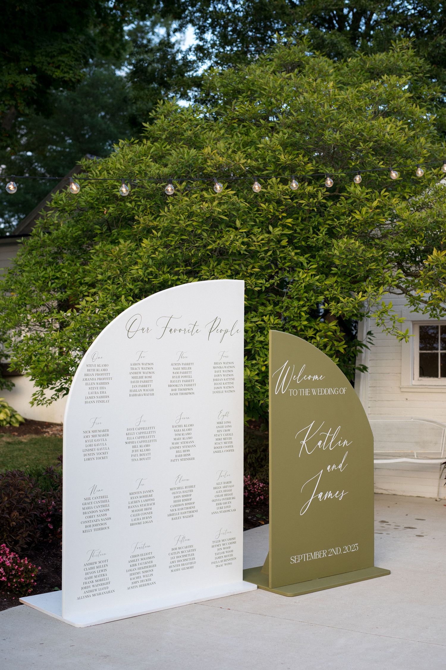 Elegant wedding seating chart display with white and olive green panels arranged outdoors under trees.
