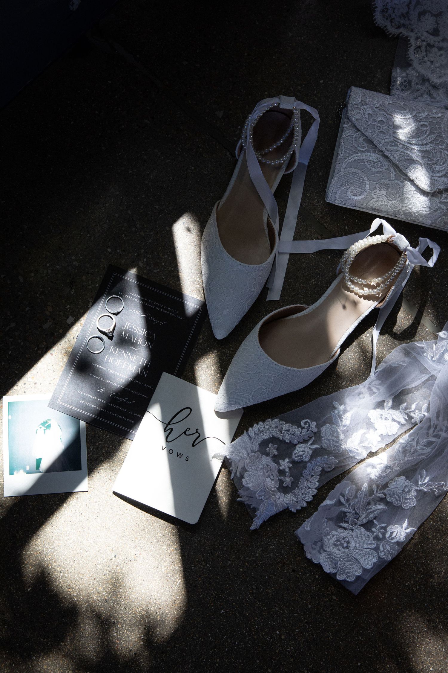 White bridal heels with pearl ankle straps displayed with wedding rings, lace accessories, and stationery in dramatic sunlight.